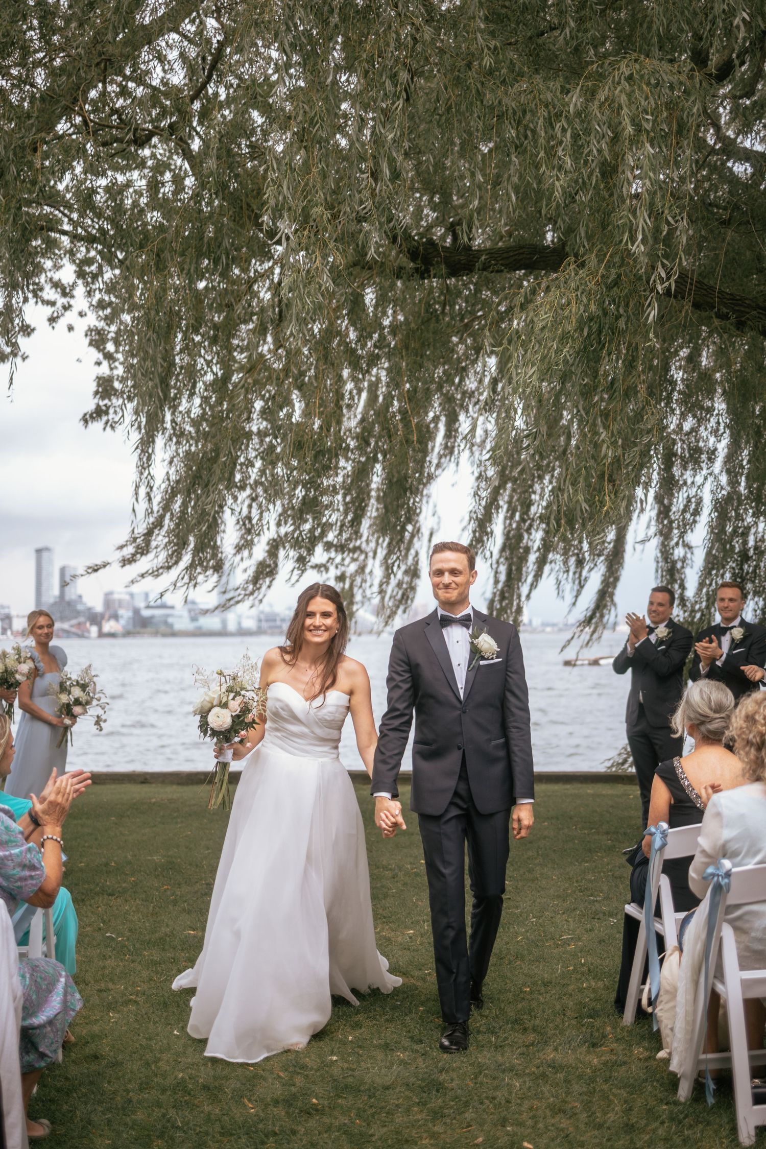 Elegant outdoor waterfront wedding ceremony with couple walking down the aisle under a weeping willow tree.