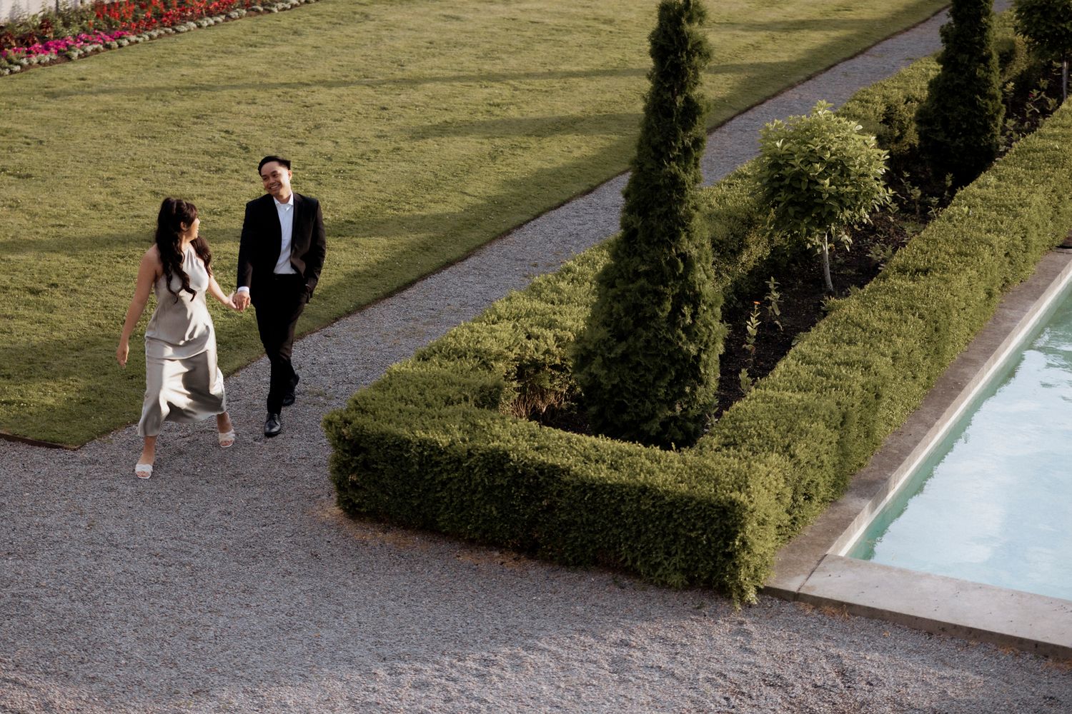 A well-dressed couple walks along a manicured garden path near formal hedges and a pool.