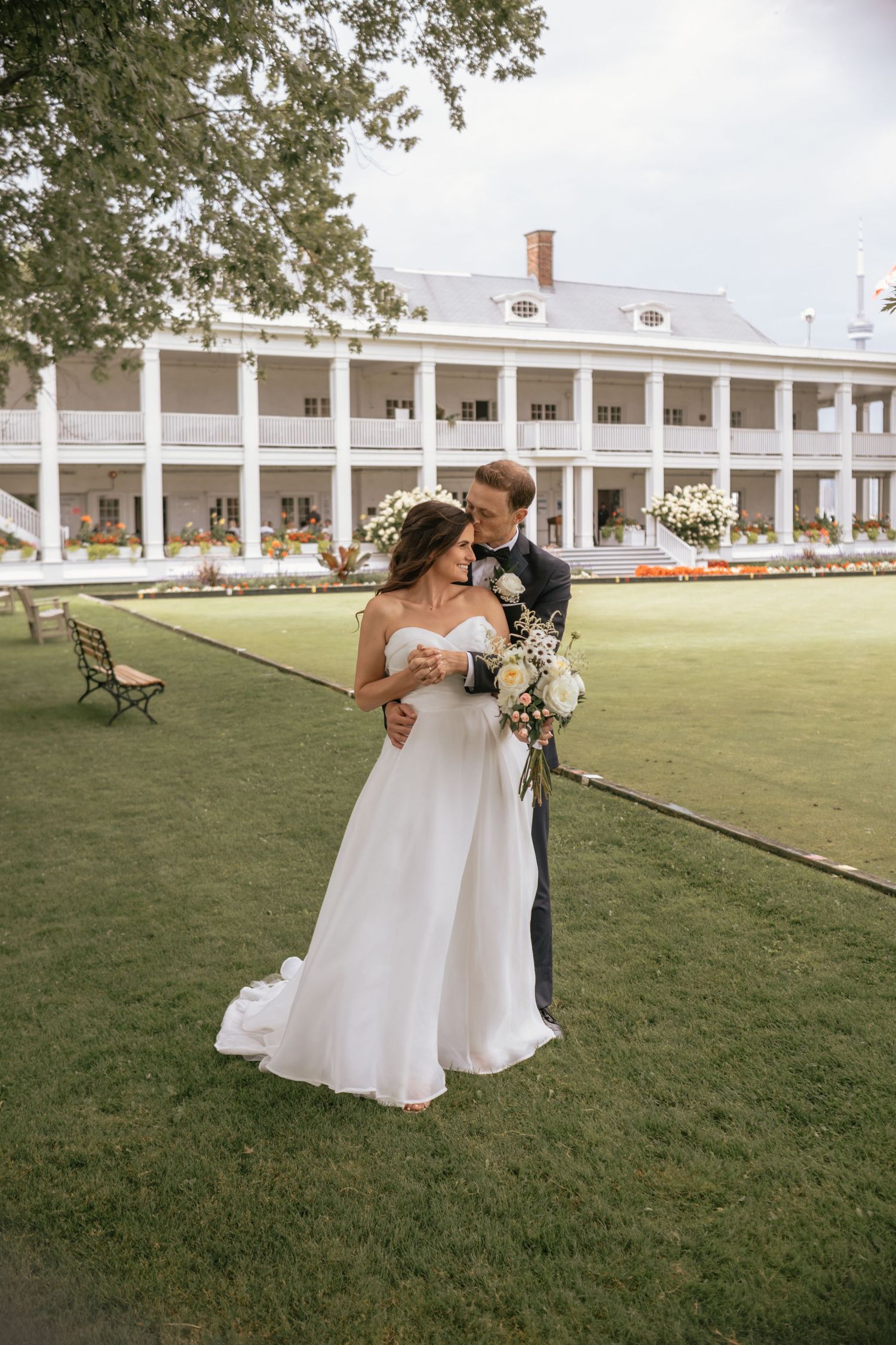 Wedding couple shares romantic moment on manicured lawn in front of elegant Southern colonial mansion with white columns.