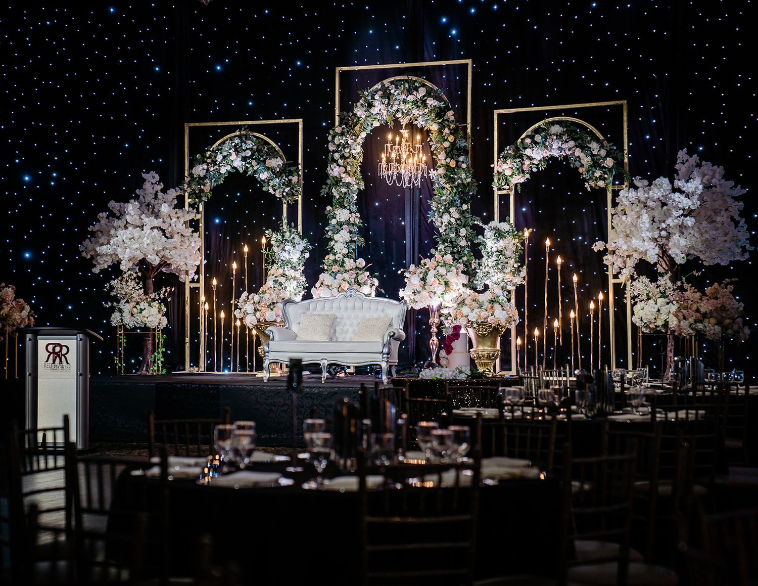 Luxurious wedding reception setup with white floral arches, candelabras and dramatic lighting against a starry backdrop.