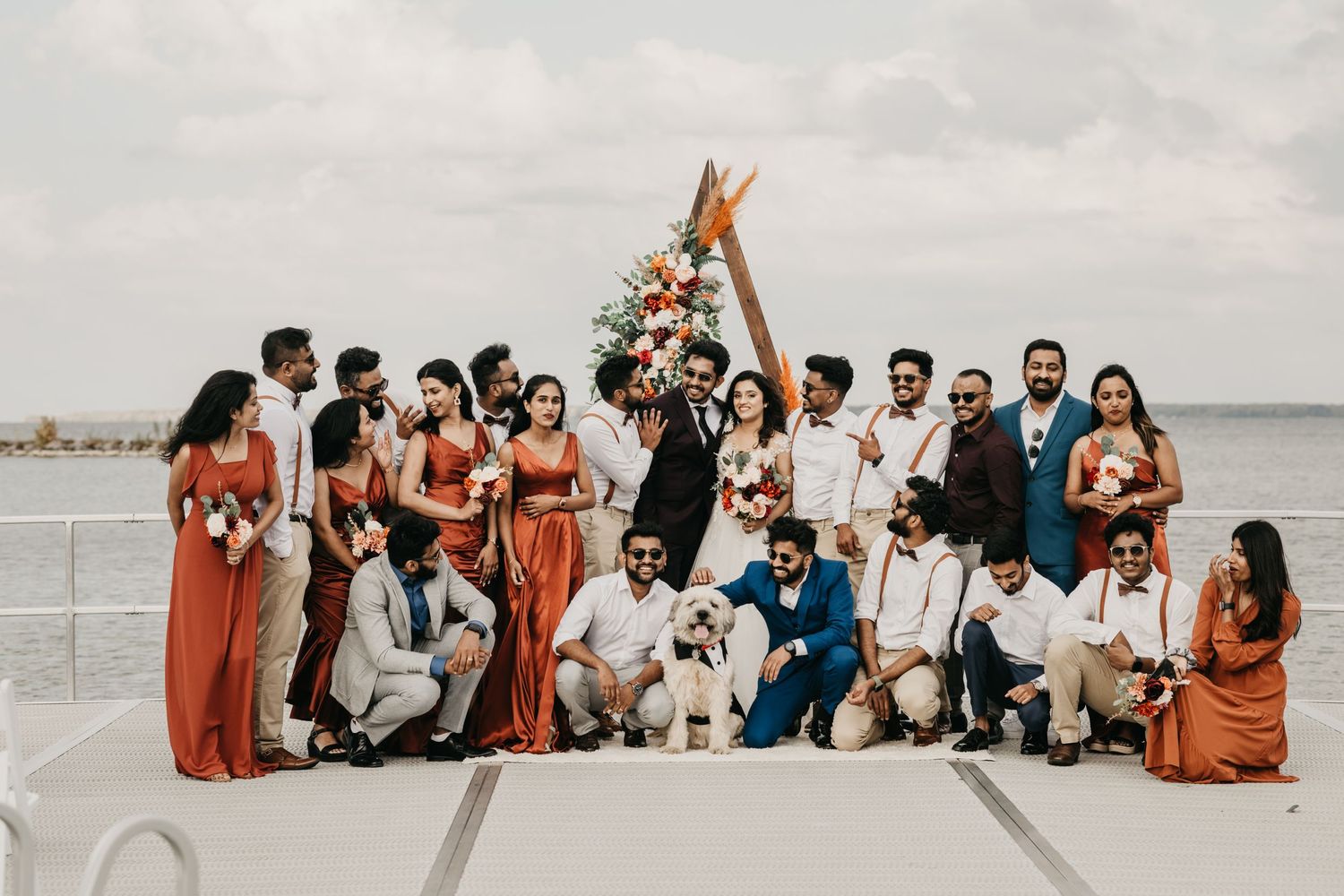 A wedding party wearing orange and navy poses together under a floral arch on a waterfront.