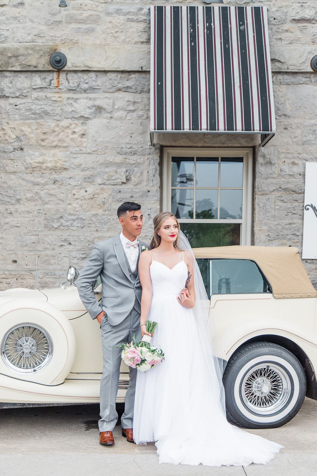 Elegant wedding photo with newlyweds posing next to a vintage cream-colored car against stone building.