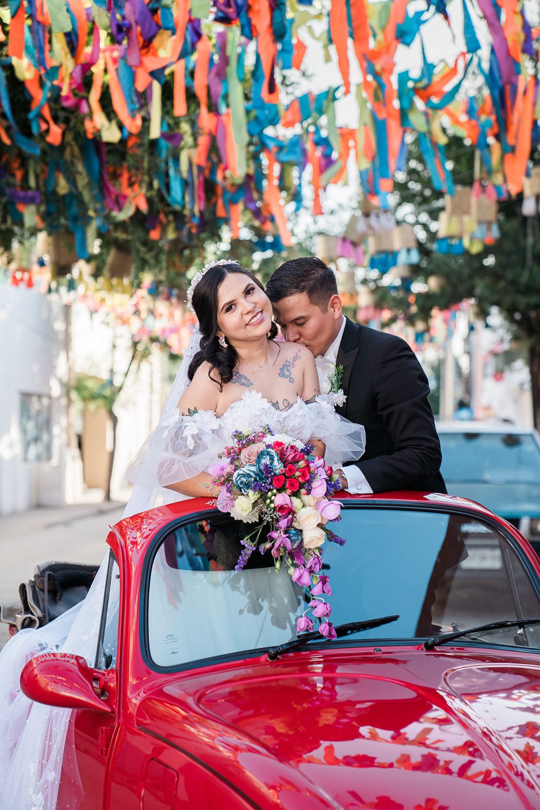 Wedding couple poses with vibrant red vintage car under colorful streamers hanging overhead.