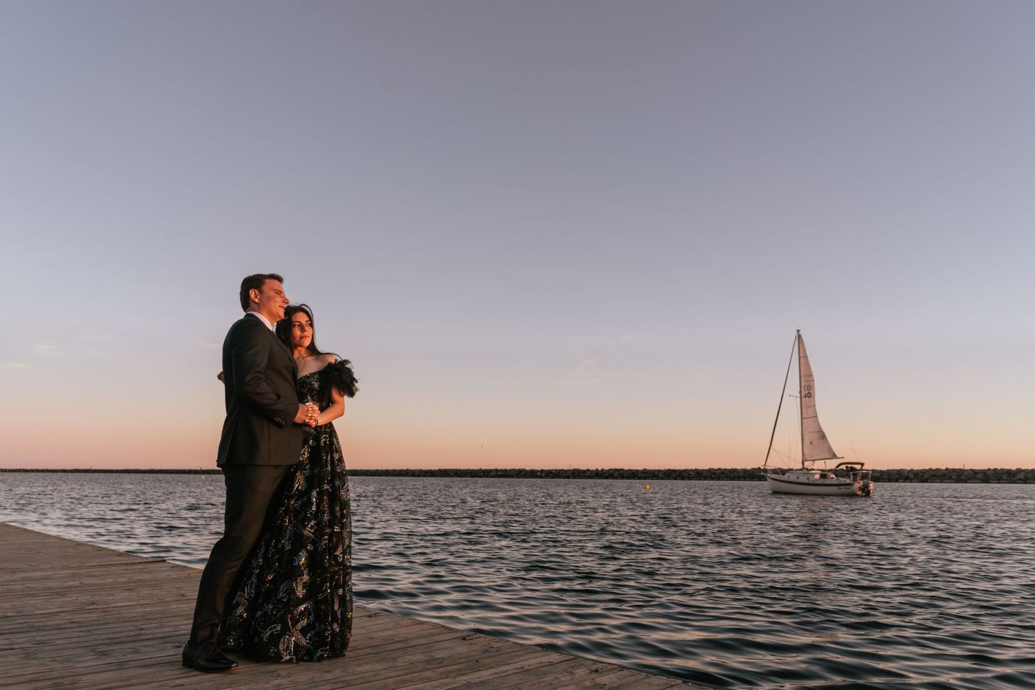 A couple in formal attire stands on a pier at sunset with a sailboat sailing in the calm waters behind them.