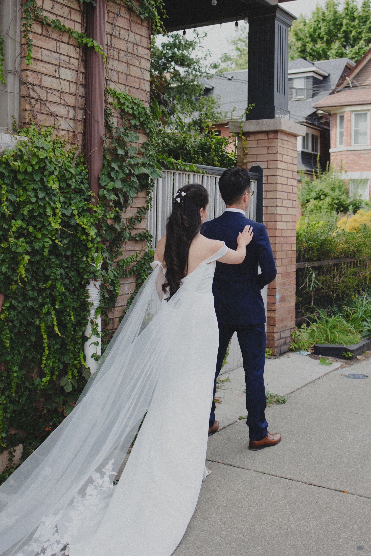 A couple in wedding attire walk down a brick-lined garden path, with the bride's long veil trailing behind them.