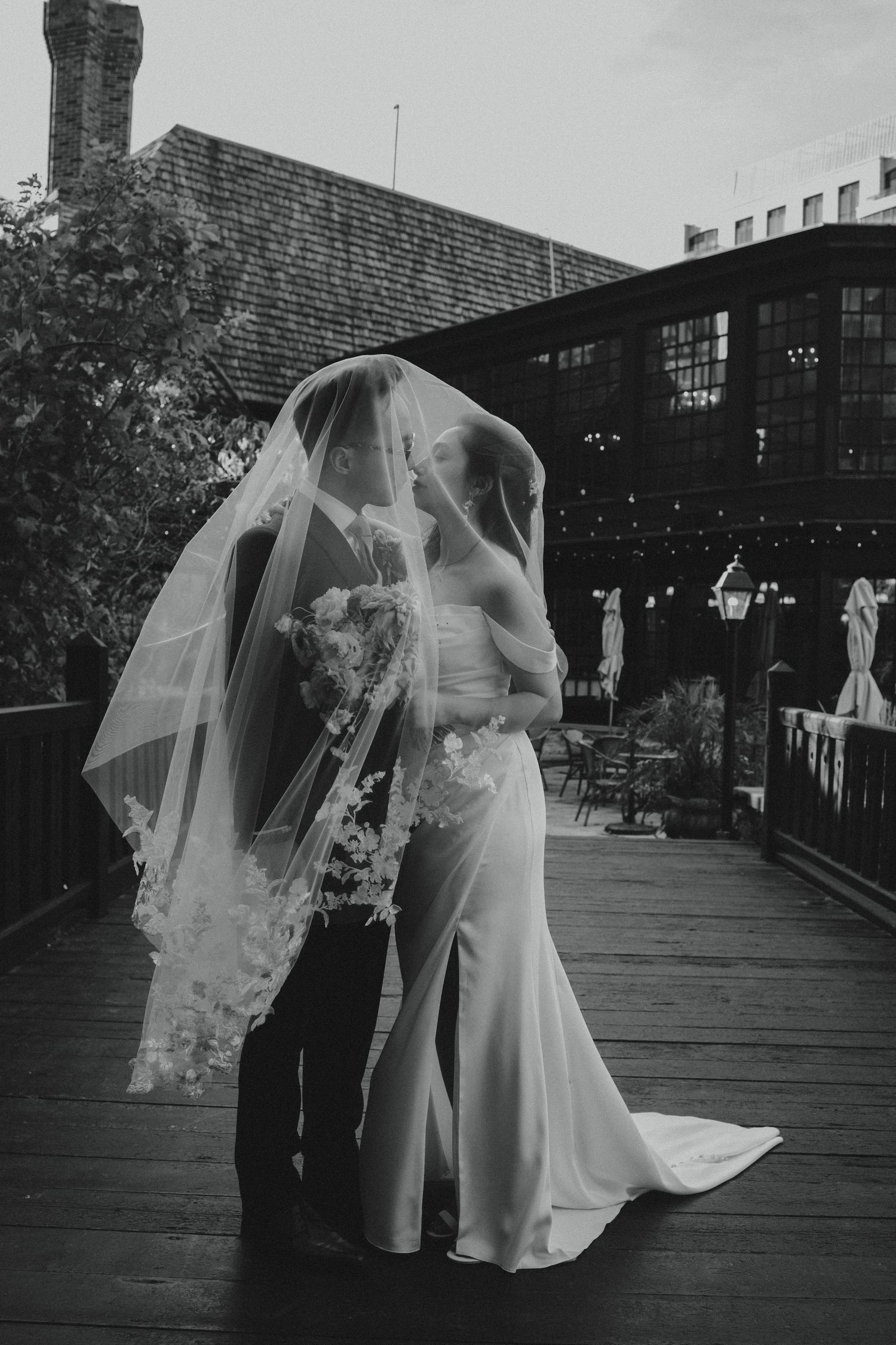 A romantic black and white wedding photo of a couple embracing under a long veil on an outdoor deck.