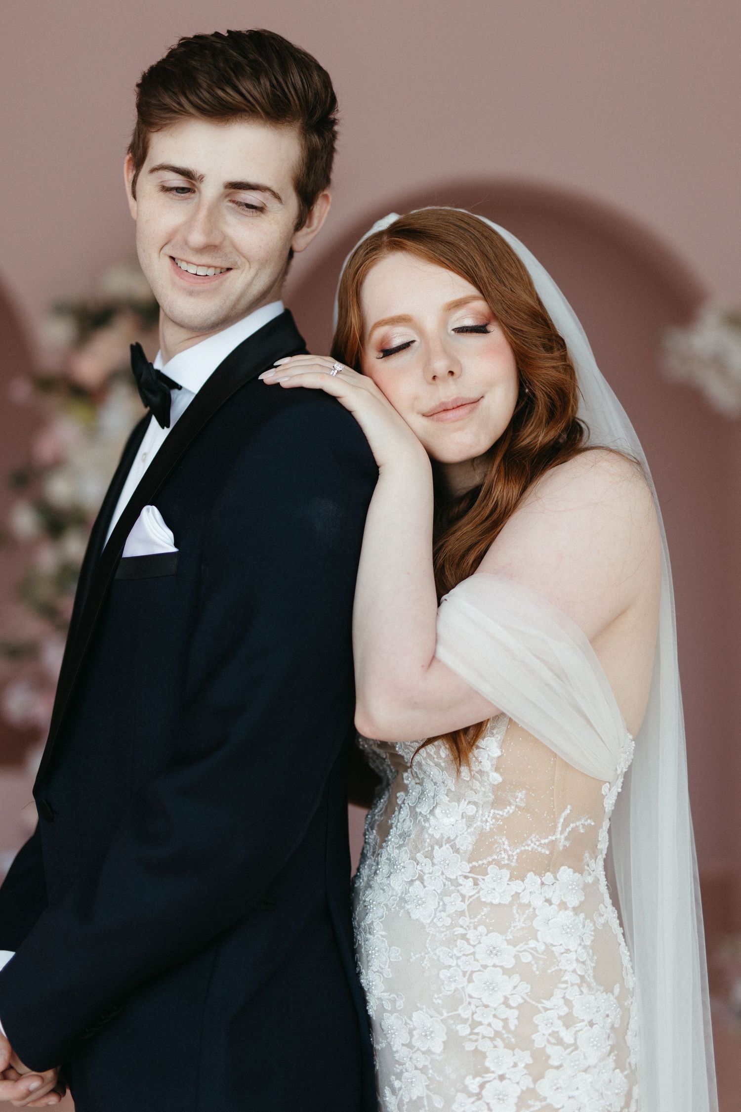 A bride in a lace wedding gown and veil leans against her groom wearing a black tuxedo with bow tie.