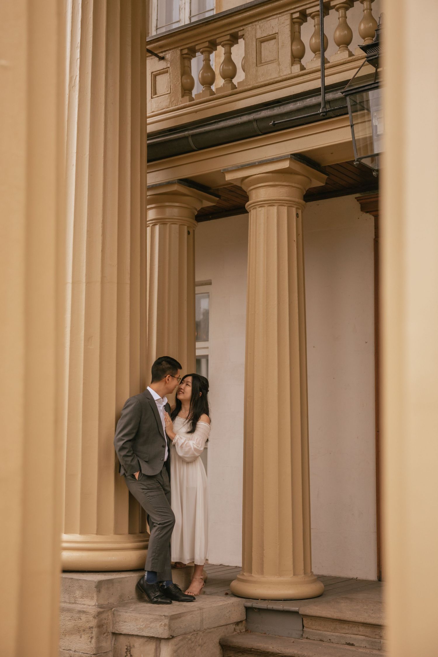 A romantic couple stands intimately between elegant cream-colored classical columns on stone steps.