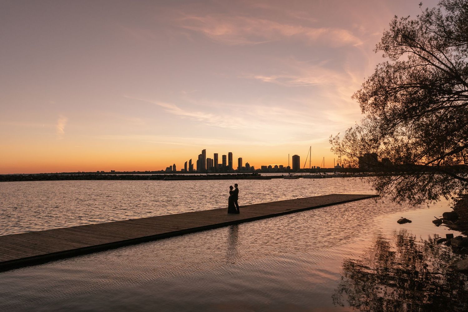 A silhouetted couple watches the sunset over the Perth city skyline from a waterfront boardwalk.