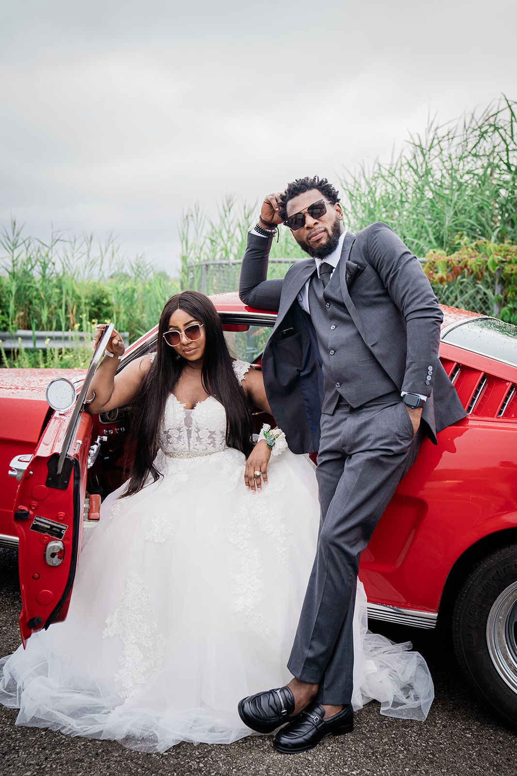 Stylish couple poses with a classic red car, wearing sunglasses and wedding attire on an overcast day.