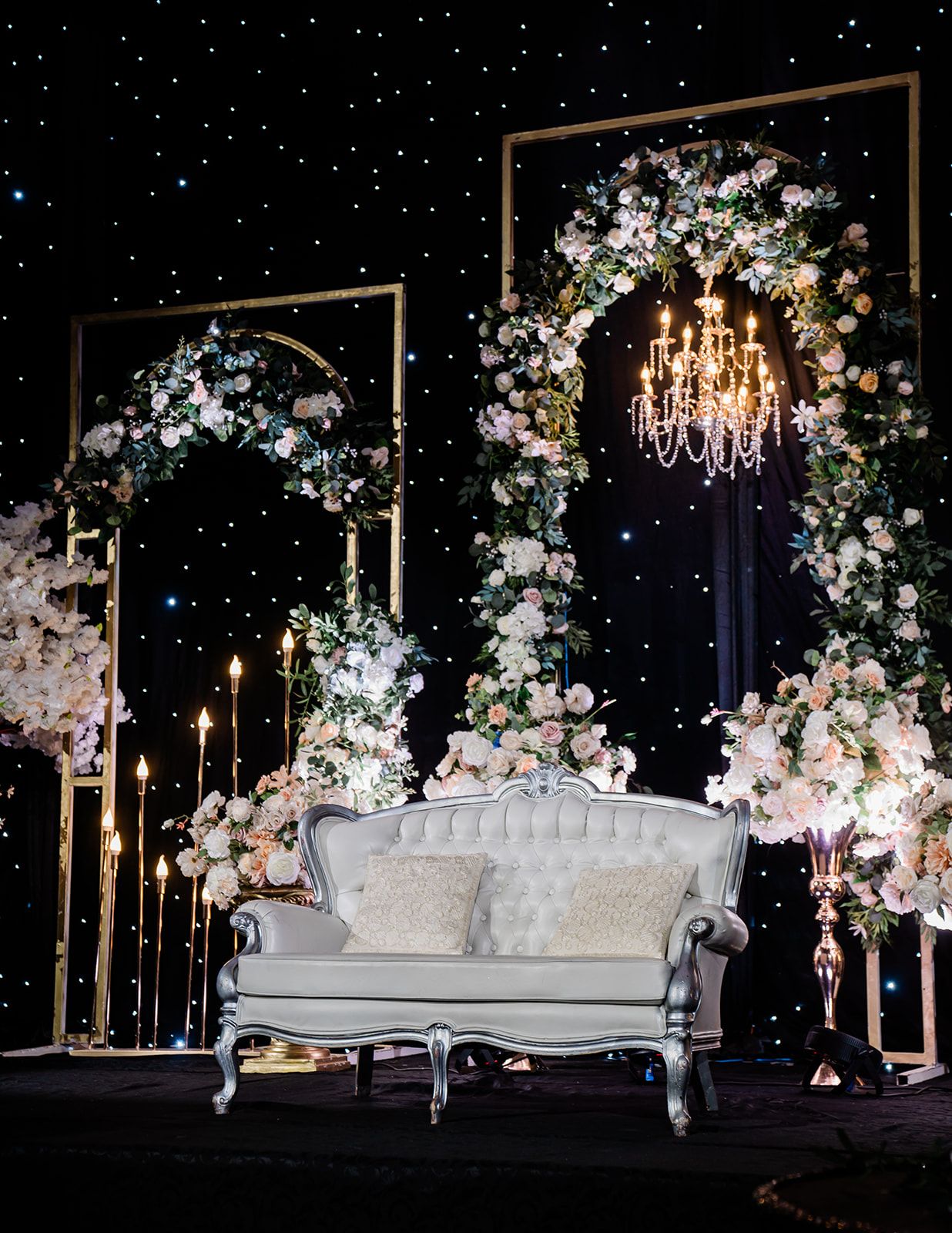 Elegant wedding seating area with white vintage sofa under floral arches and crystal chandelier against starry backdrop.
