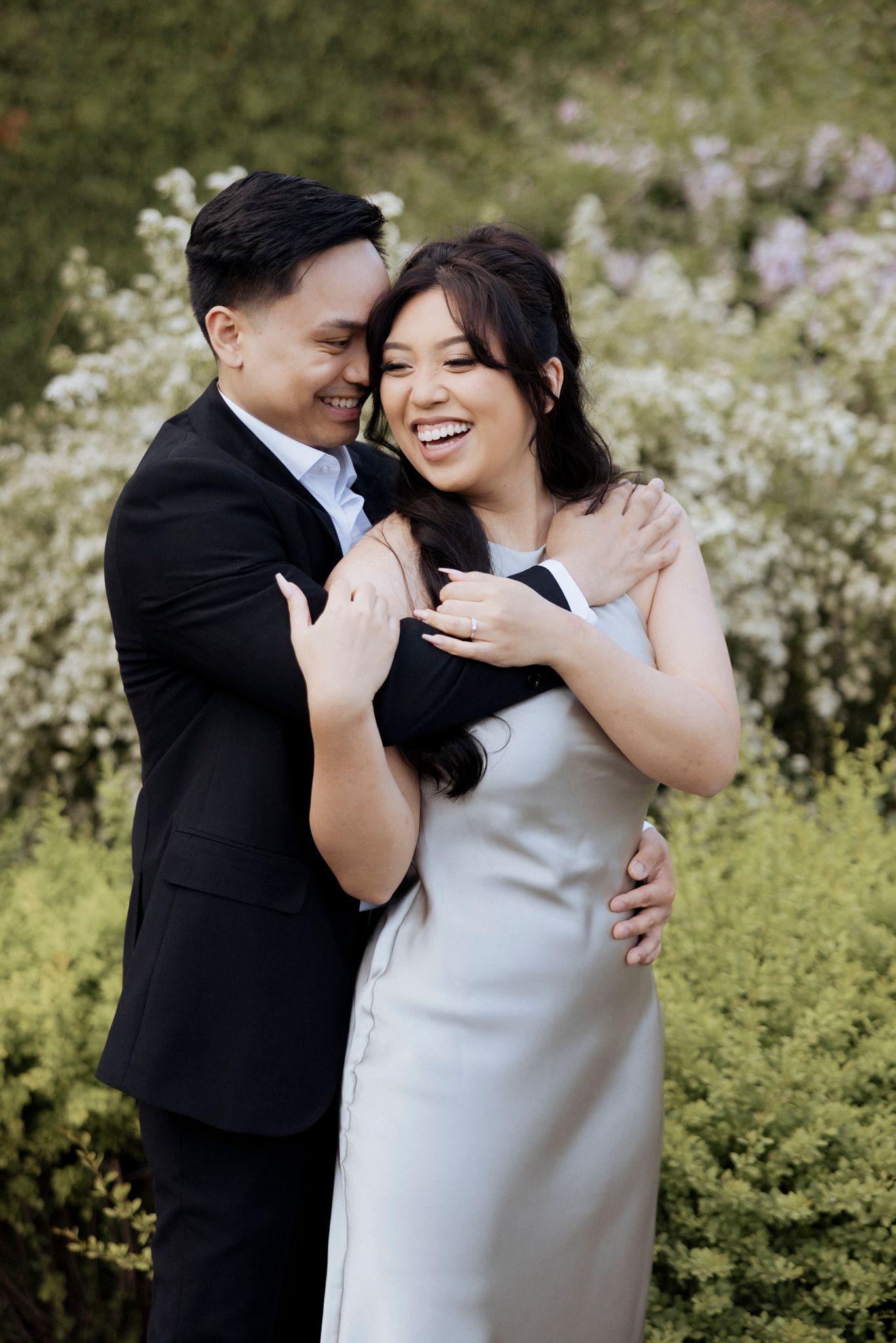 A joyful couple laughs together during their outdoor engagement photoshoot surrounded by flowers.