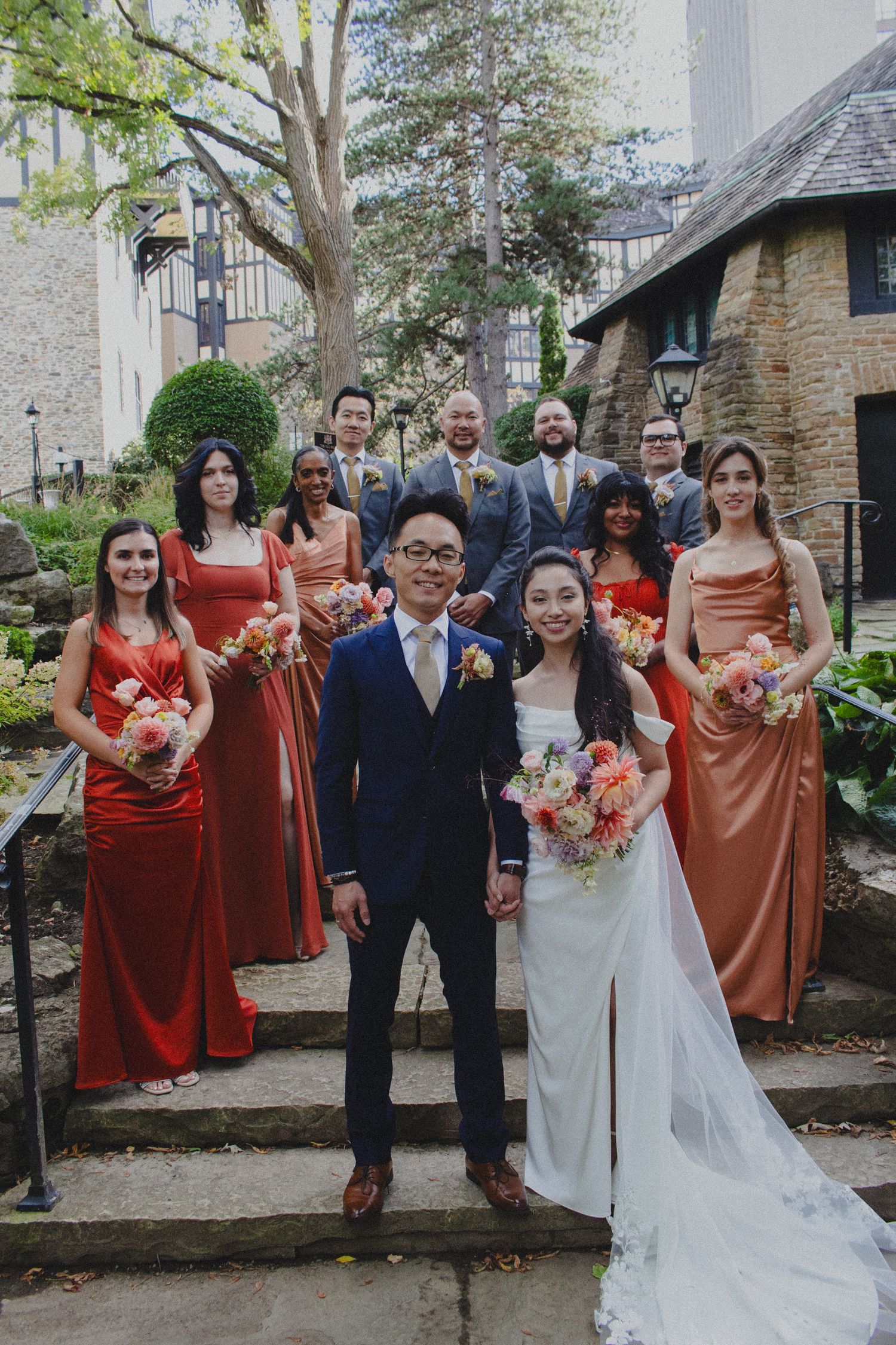 Wedding party in rust-colored bridesmaid dresses and navy suit poses on stone steps of garden courtyard.