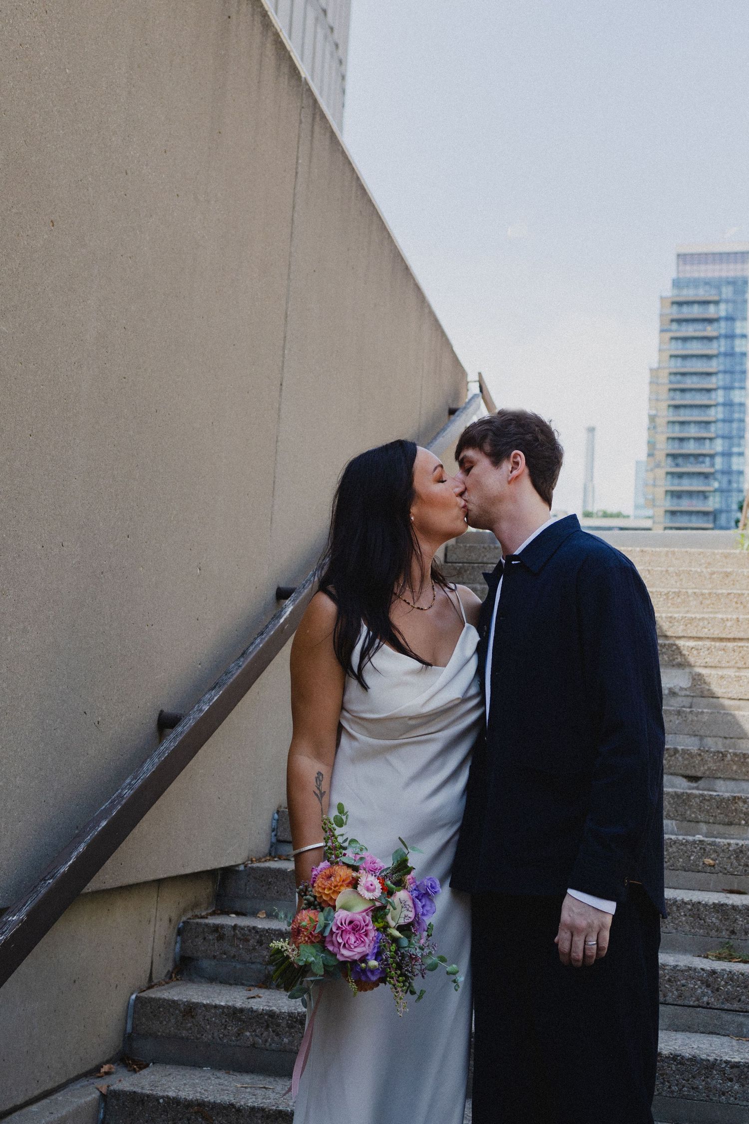 Couple sharing a romantic moment on urban concrete steps, bride in silk dress holding colorful floral bouquet.