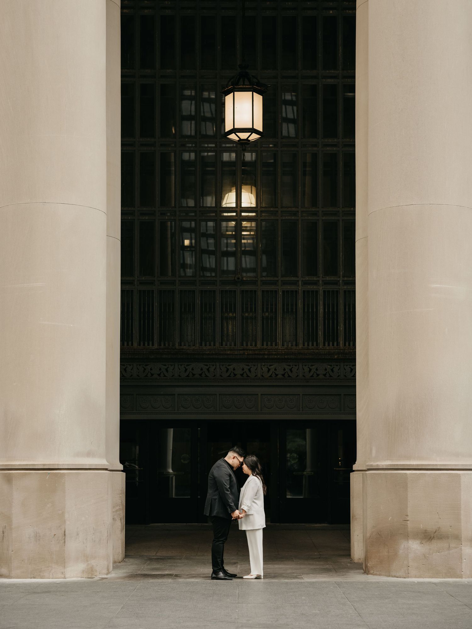 A couple shares a romantic moment between grand white stone columns with ornate architectural lighting above.