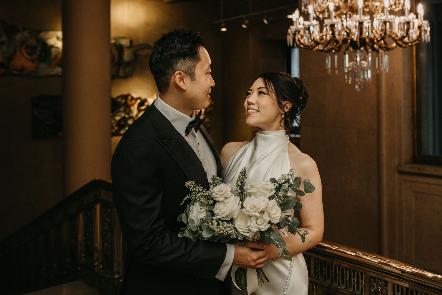 A romantic moment captured in warm lighting with an ornate chandelier as a couple poses with ivory floral bouquet indoors.