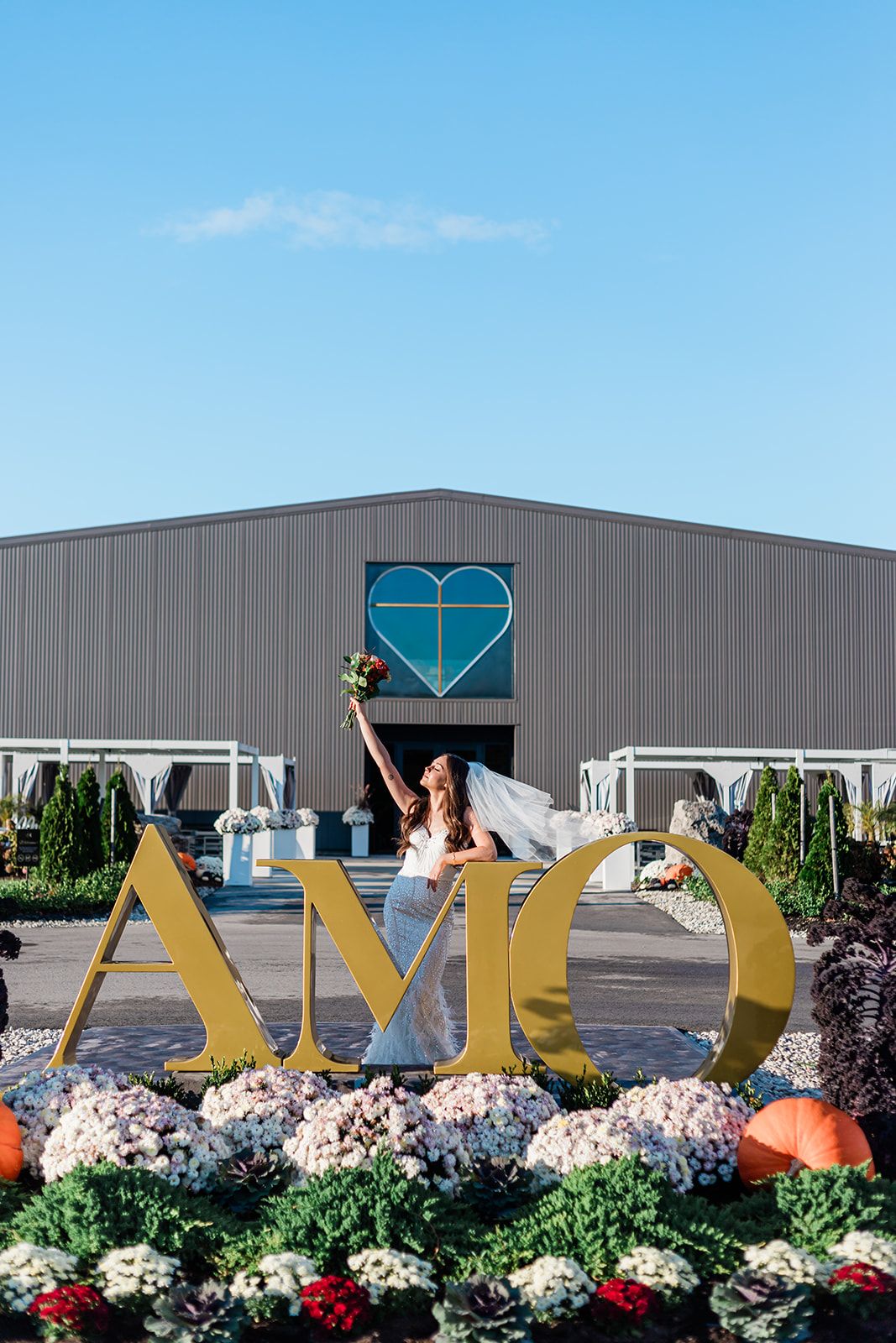 A person poses excitedly by large gold AMD letters surrounded by flowers and pumpkins outside a building with a heart logo.