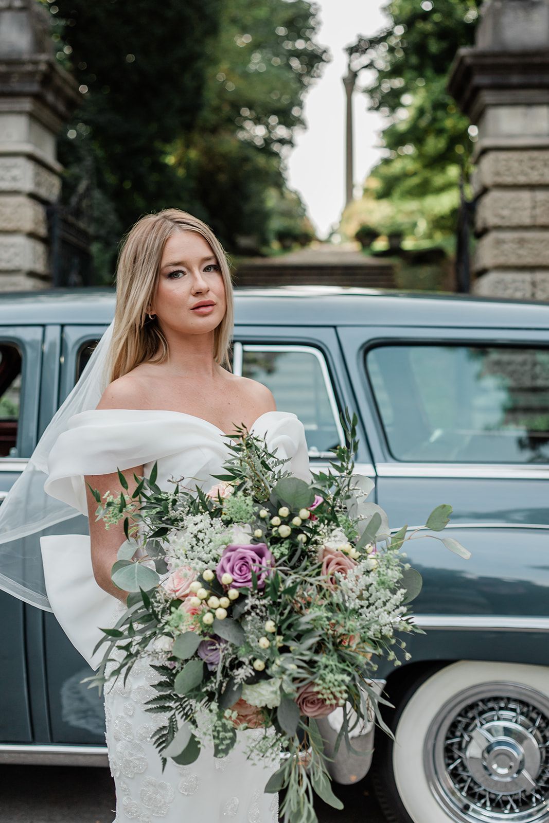 Elegant bride in white off-shoulder gown holds lush eucalyptus and rose bouquet beside vintage car at stone entrance.