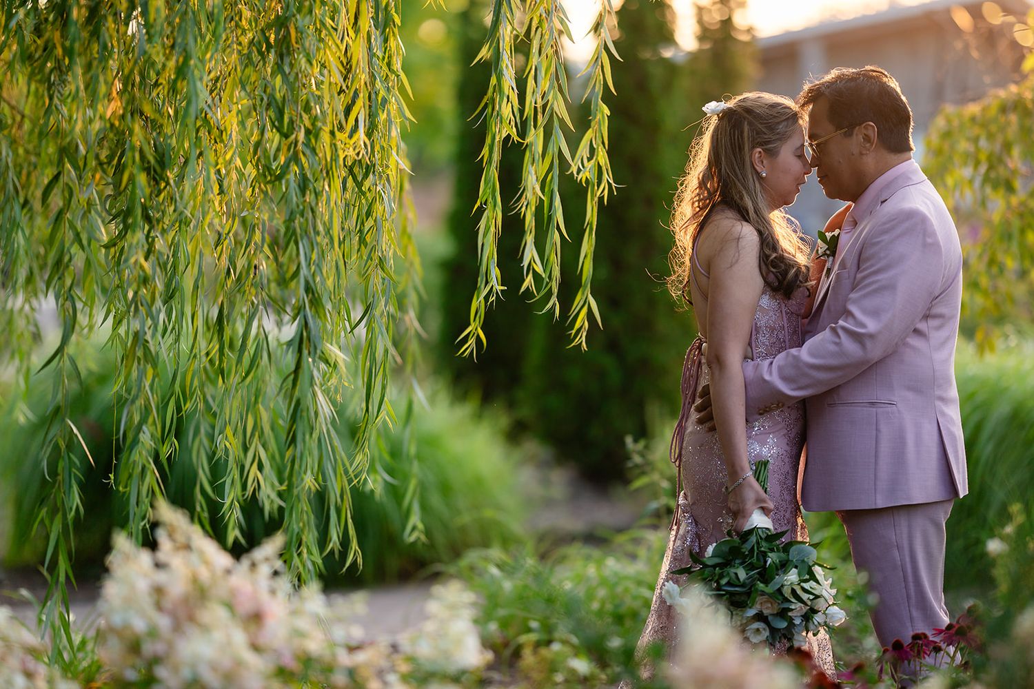A wedding couple shares an intimate moment under weeping willow trees at sunset.