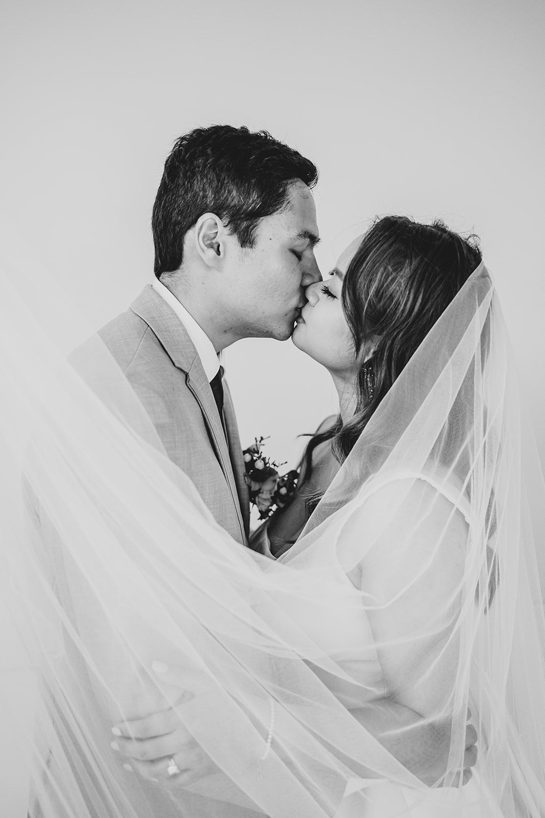 A romantic black and white wedding photo of a couple kissing under a flowing white veil.