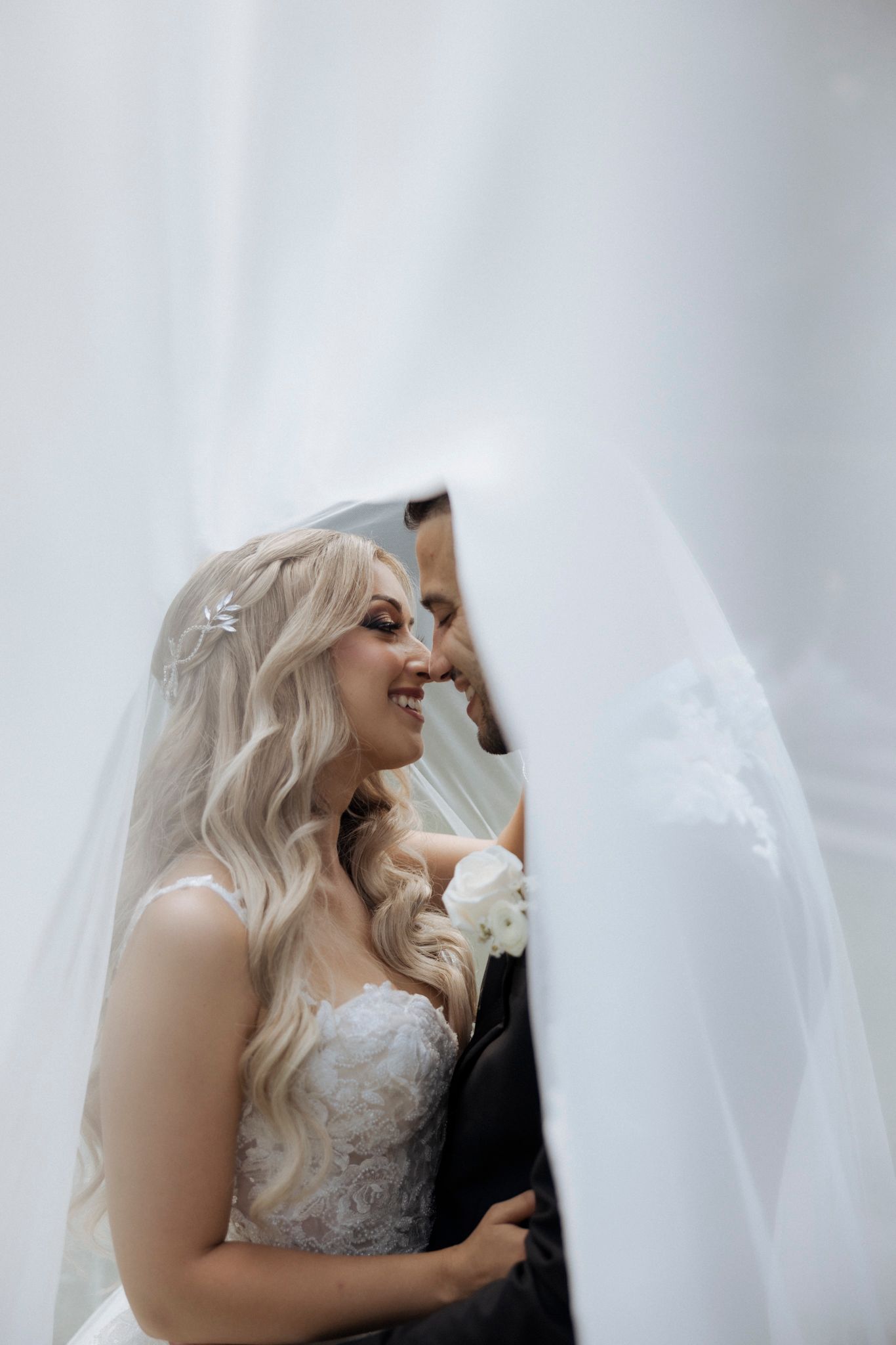 Bride in white lace dress smiling through wedding veil.