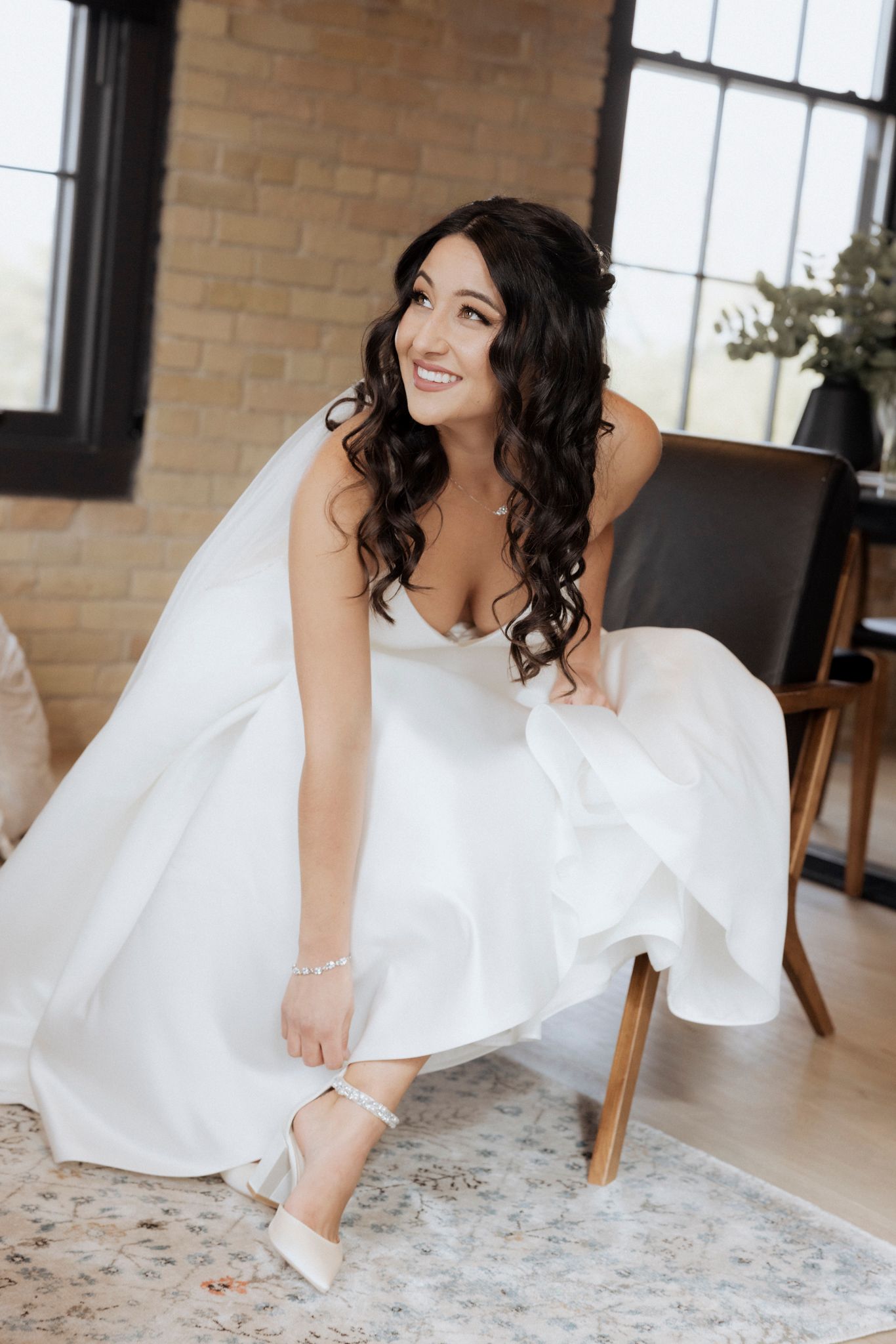A bride in a flowing white gown adjusts her shoe while seated in an elegant room with brick walls.
