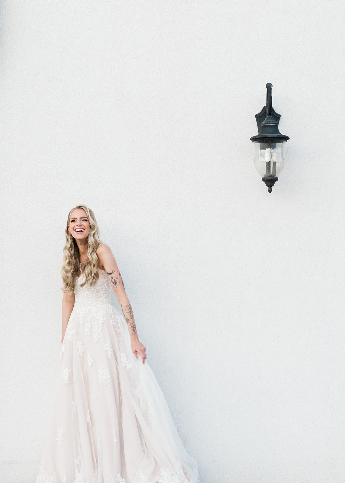 A bride in an elegant white wedding dress poses against a white wall with a vintage lantern.