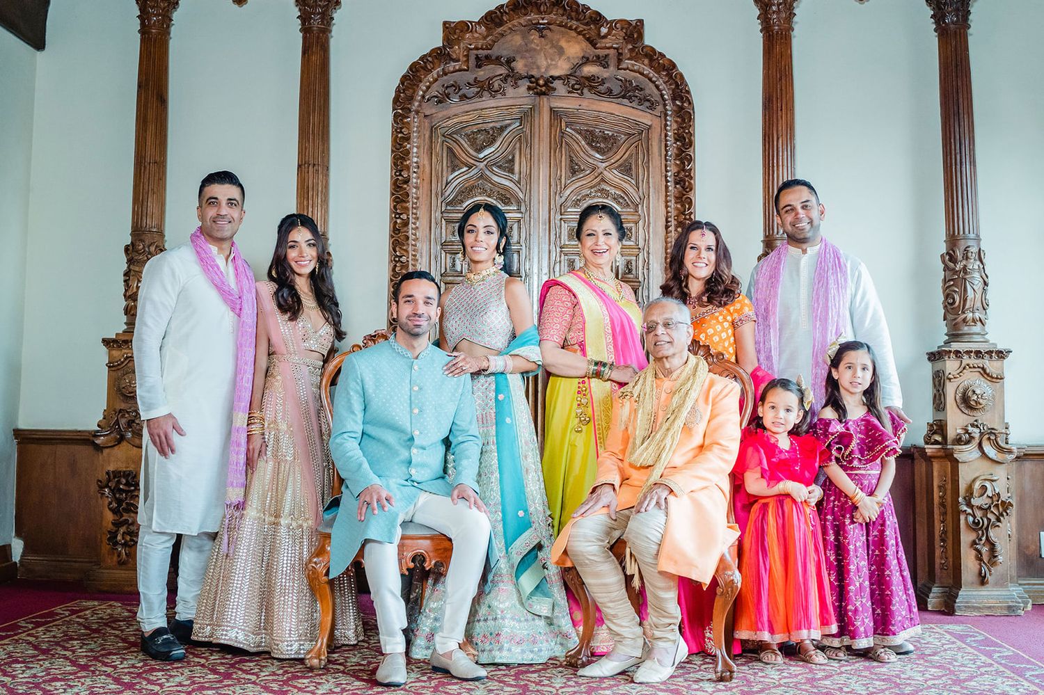 Colorful group photo in traditional Indian attire posed in front of ornate carved wooden decor.