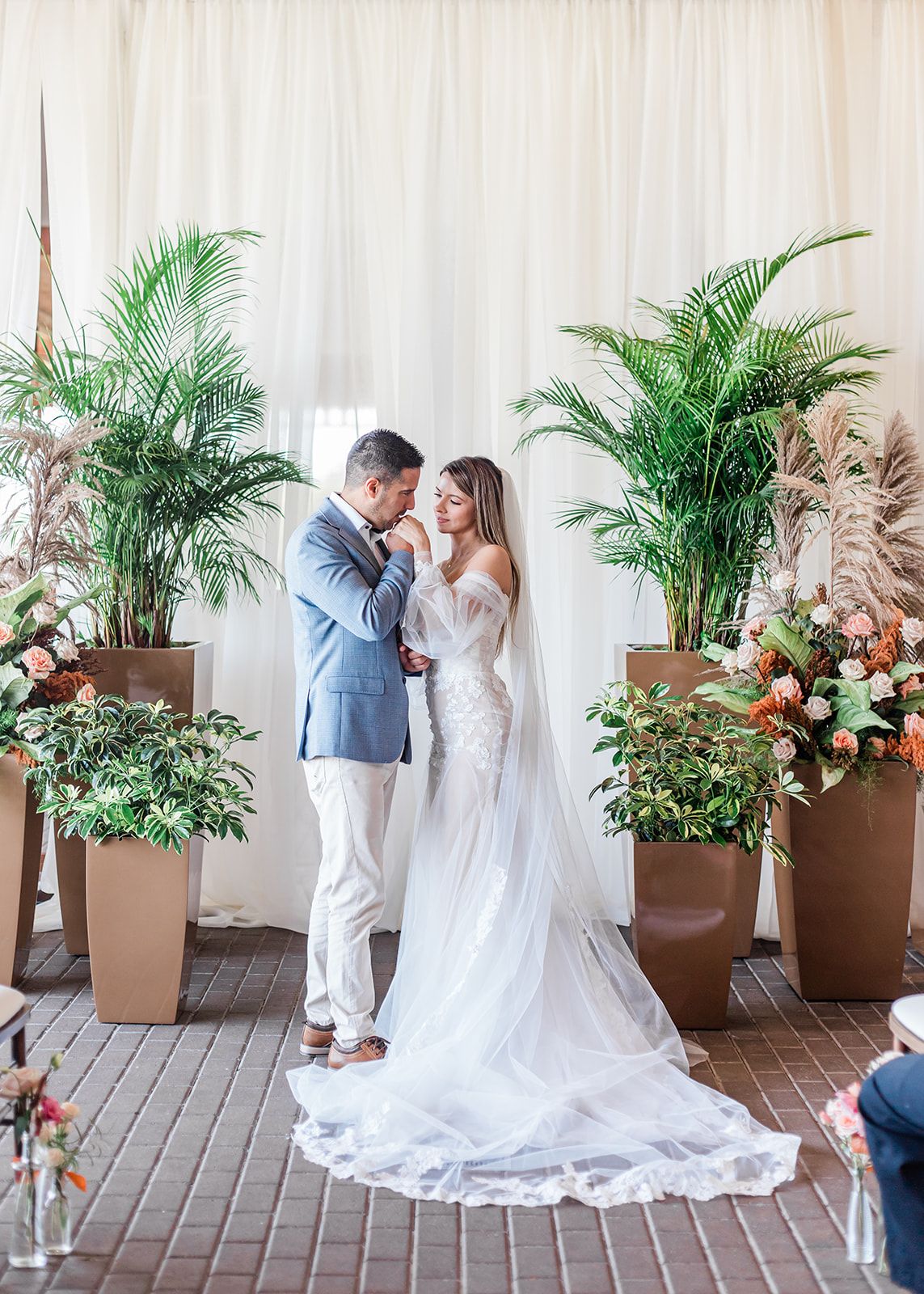 A romantic wedding ceremony setup with tropical palms and floral arrangements framing a couple in white.