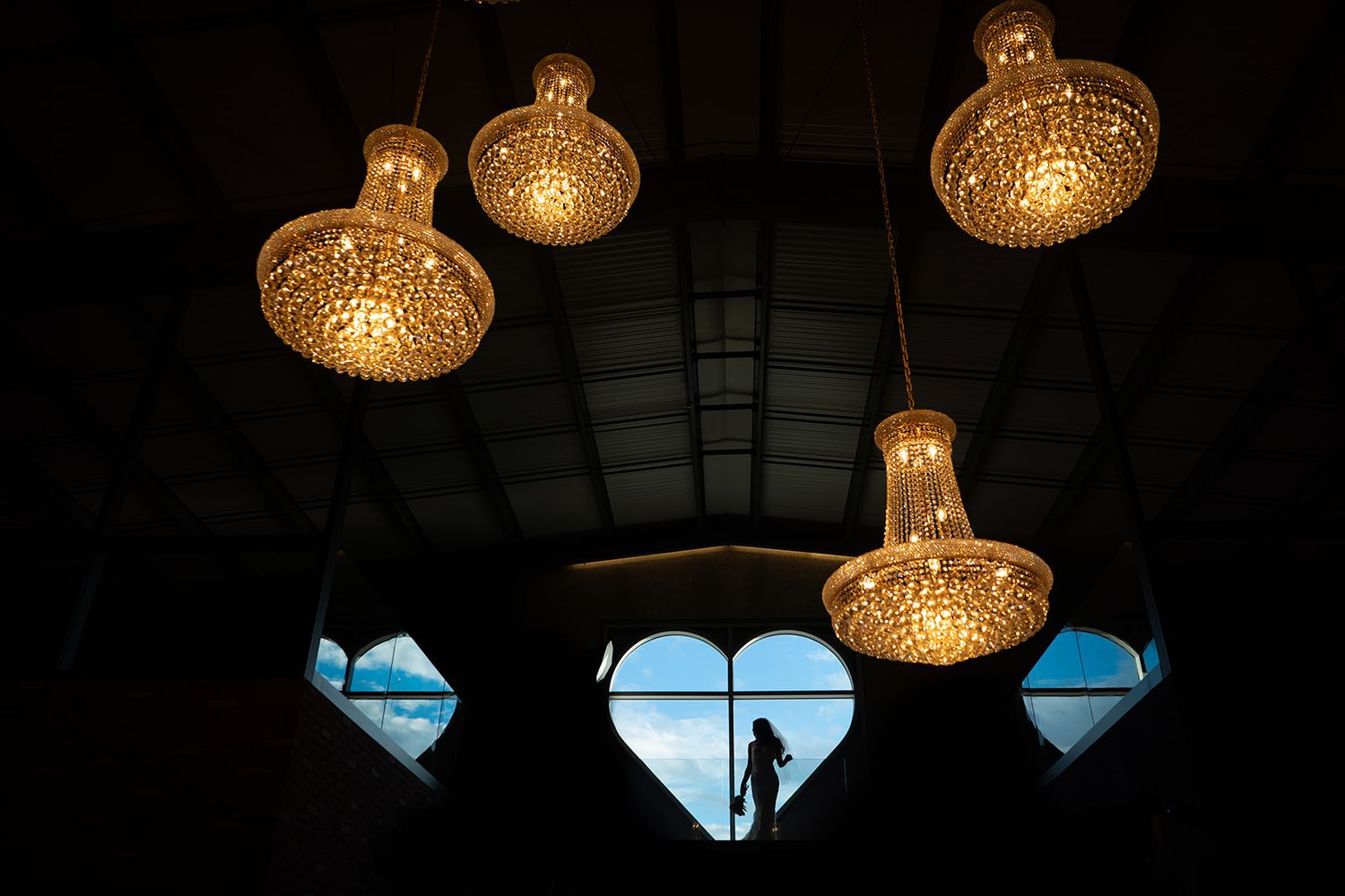 Crystal chandeliers illuminate a dark interior with heart-shaped windows, silhouetting a figure against a blue sky.