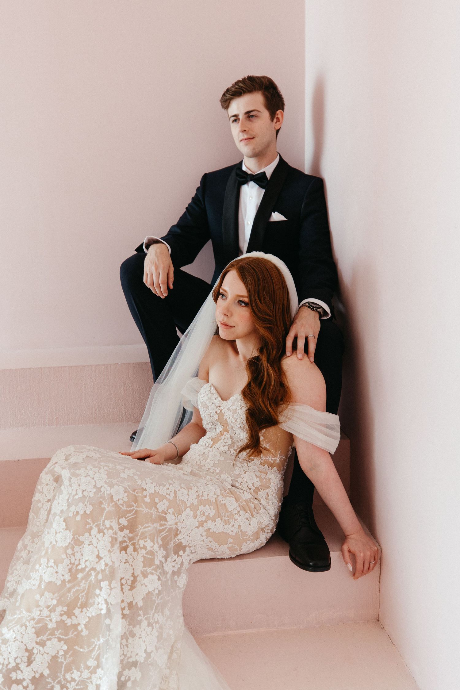 A bride in a lace gown and groom in classic tuxedo pose together on pink stairs during their wedding photoshoot.