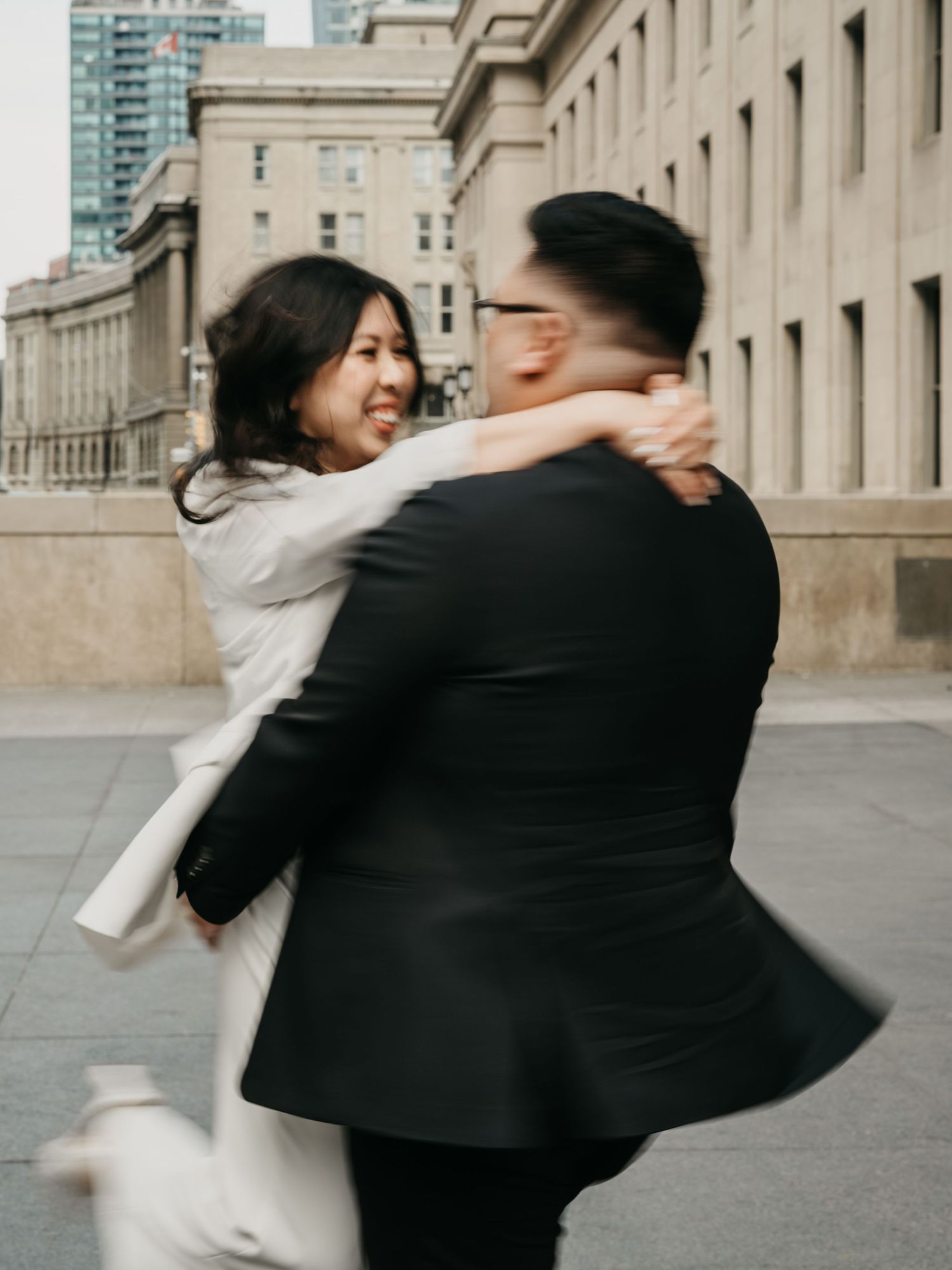 A couple embraces and spins joyfully outside a classical stone building with modern skyscrapers in the background.