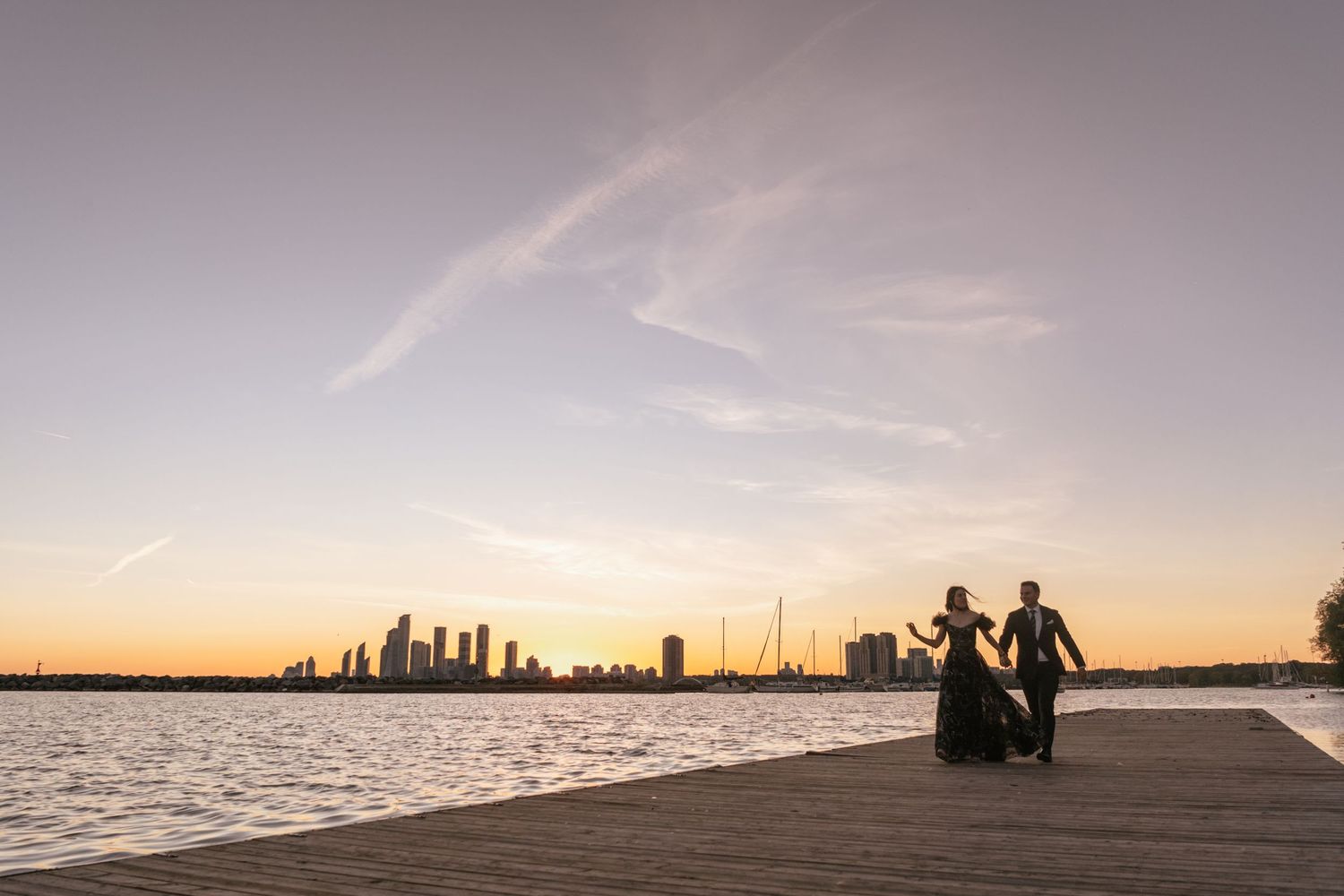 Two silhouettes holding hands on a waterfront pier at sunset with Toronto city skyline in the background.