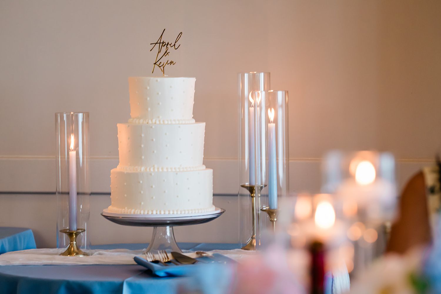 Elegant three-tier white wedding cake displayed on table with glass candle holders and soft lighting.
