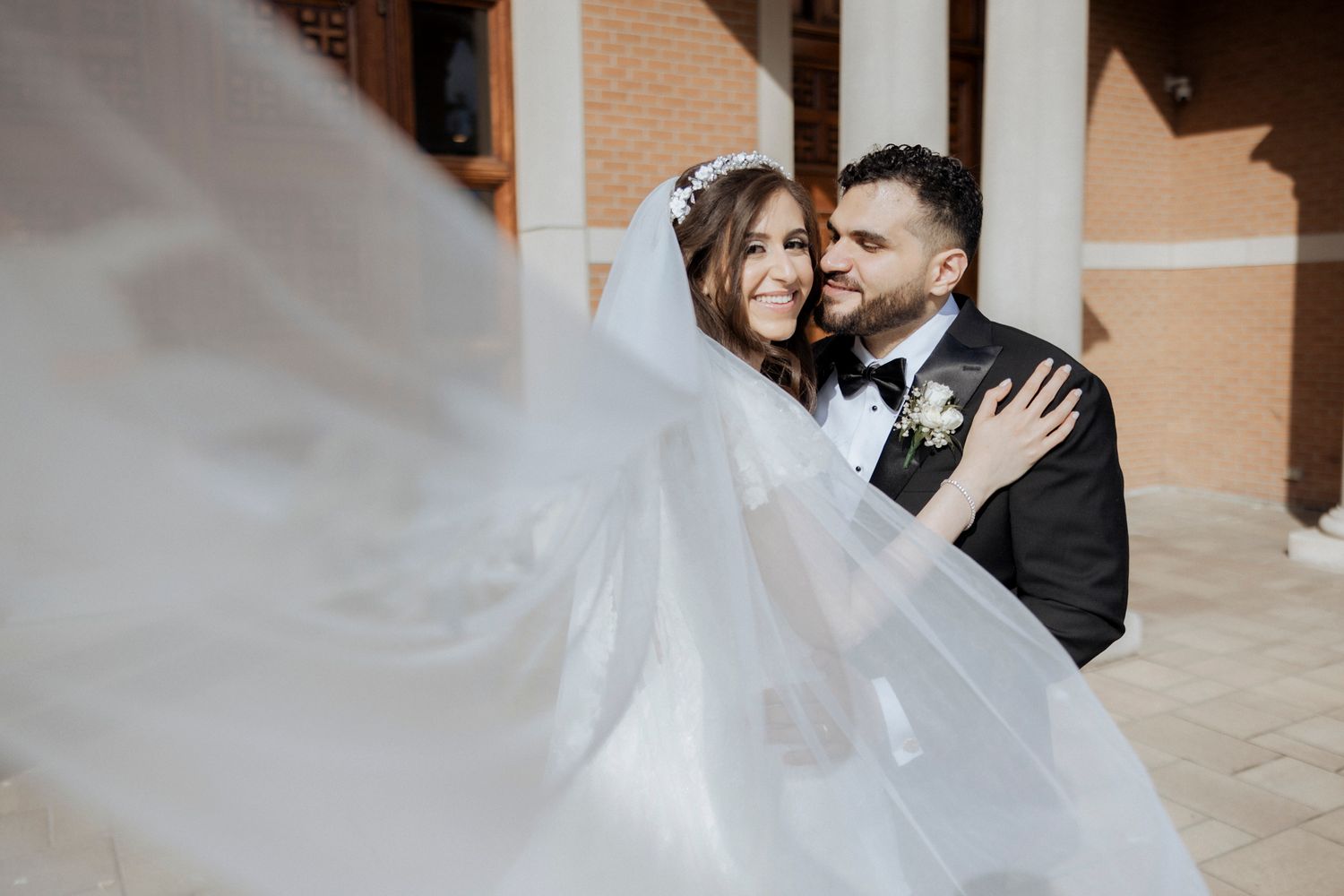 Wedding couple sharing intimate moment as bride's veil flows in wind against brick facade.