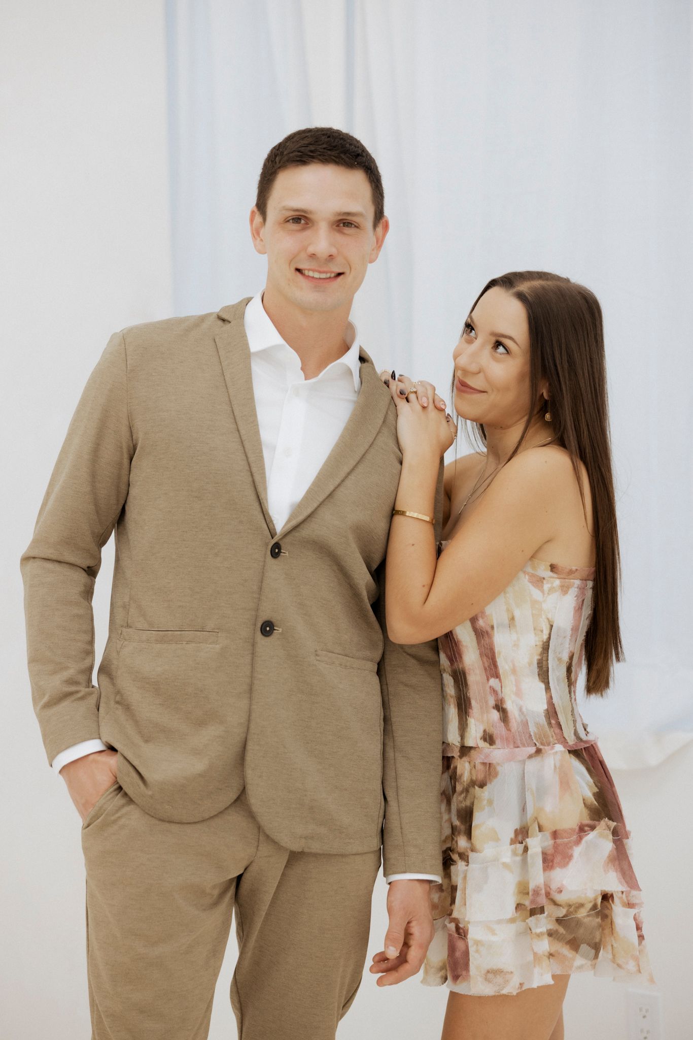A couple poses together against a white backdrop - he in a tan suit, she in a floral print summer dress.