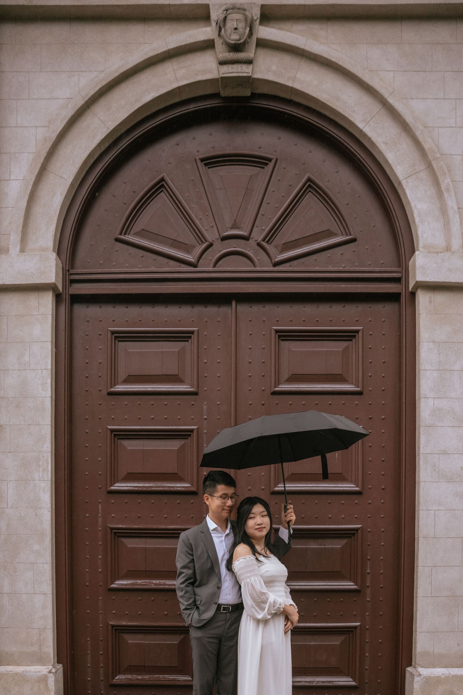 A couple stands together holding an umbrella in front of a large brown arched door with decorative panels.