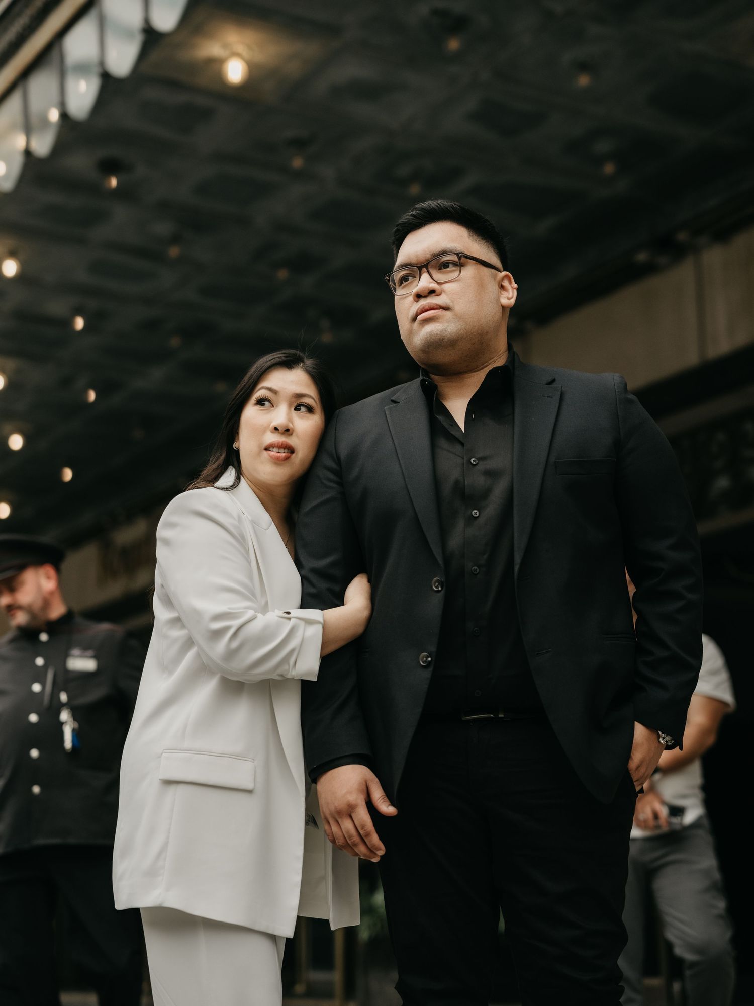A couple stands together indoors under dim lighting, with one wearing a black suit and one in a light blazer.