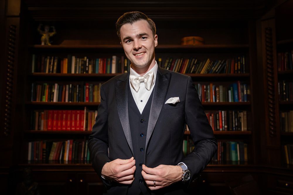 Someone in a black tuxedo and bow tie smiles while standing in front of a library bookshelf.