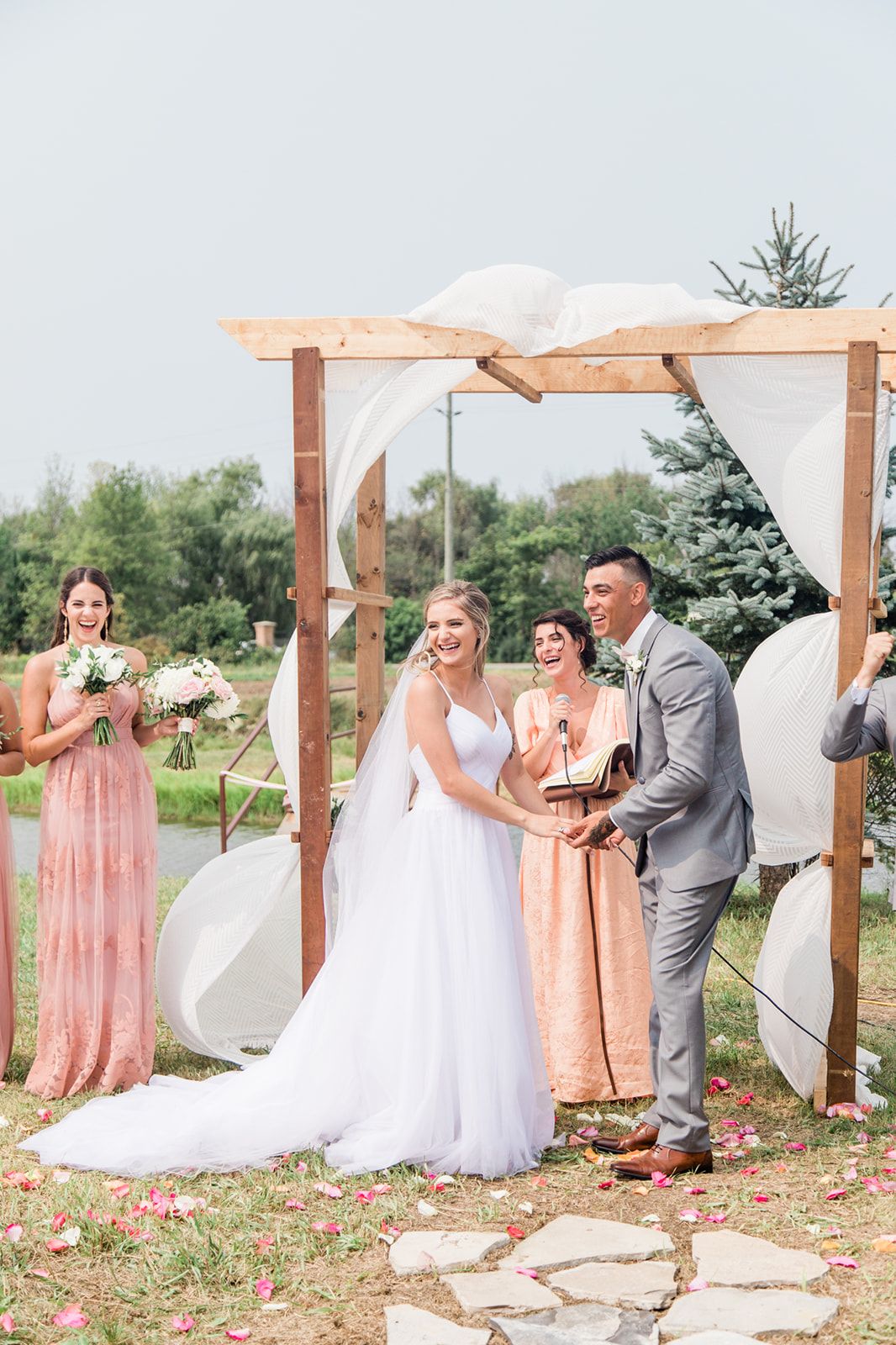 A wedding ceremony takes place under a rustic wooden pergola with bridesmaids in peach dresses and scattered rose petals.