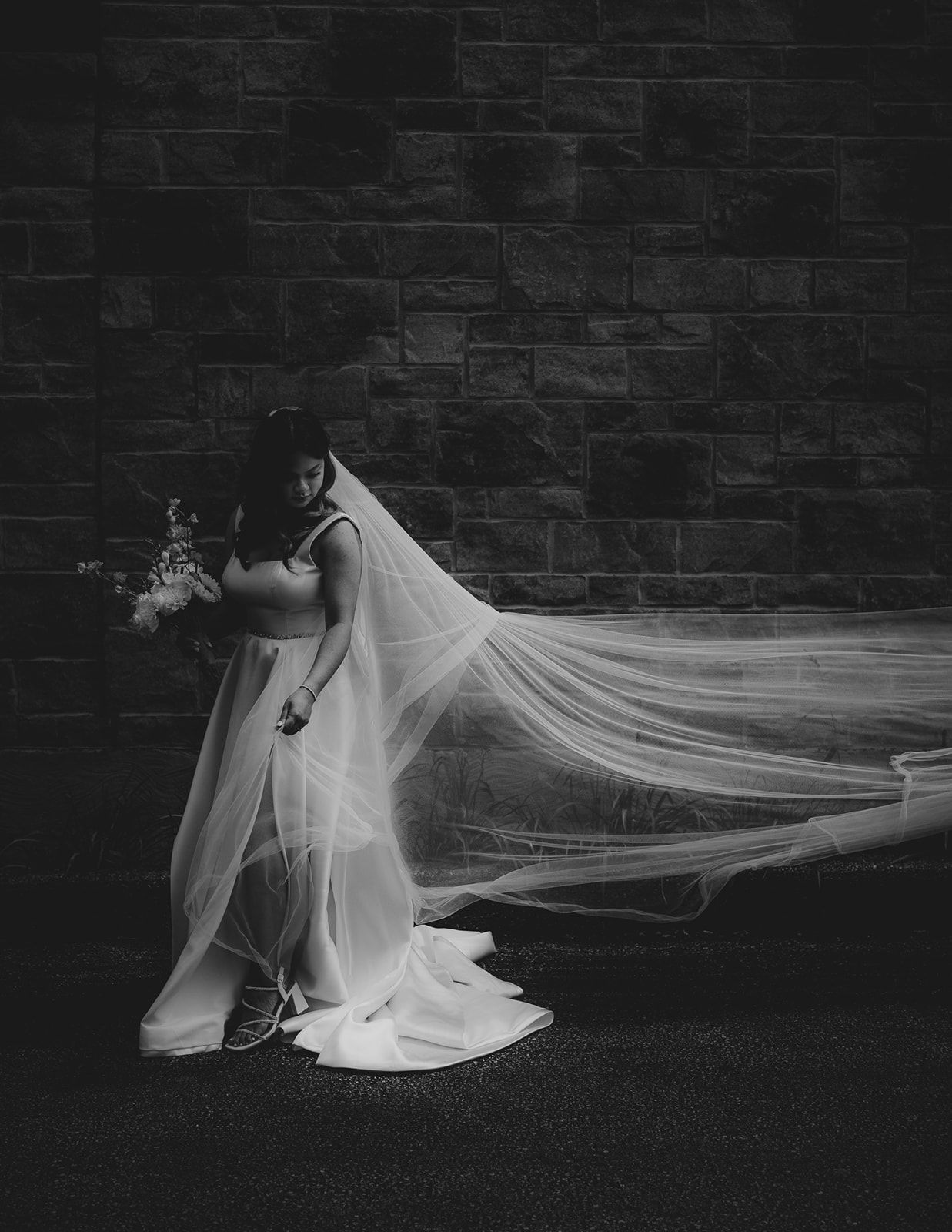 A dramatic black and white bridal portrait showing a flowing veil against a brick wall.