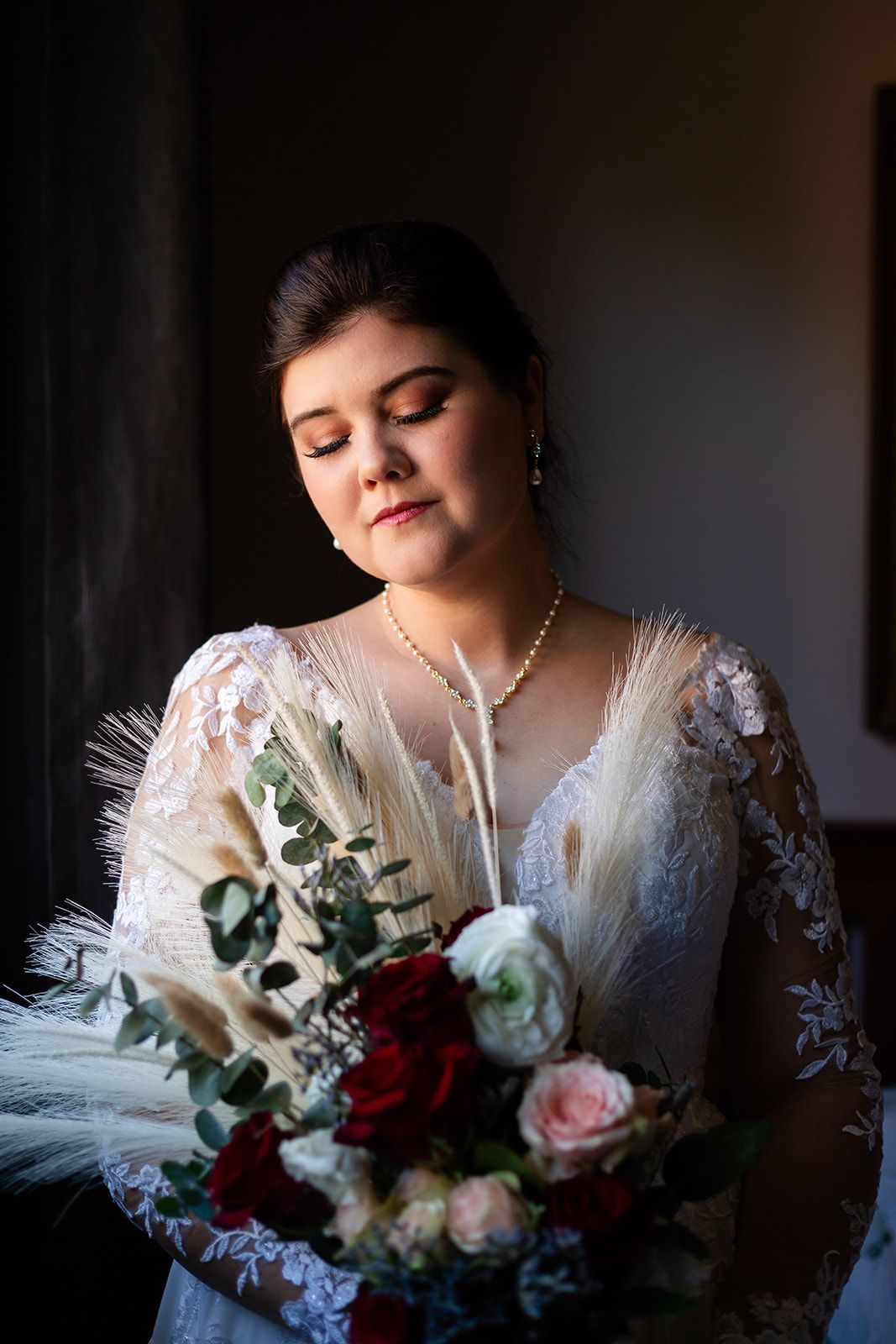 Romantic bridal portrait with delicate lace details and a mixed bouquet of red and pink roses in moody lighting.