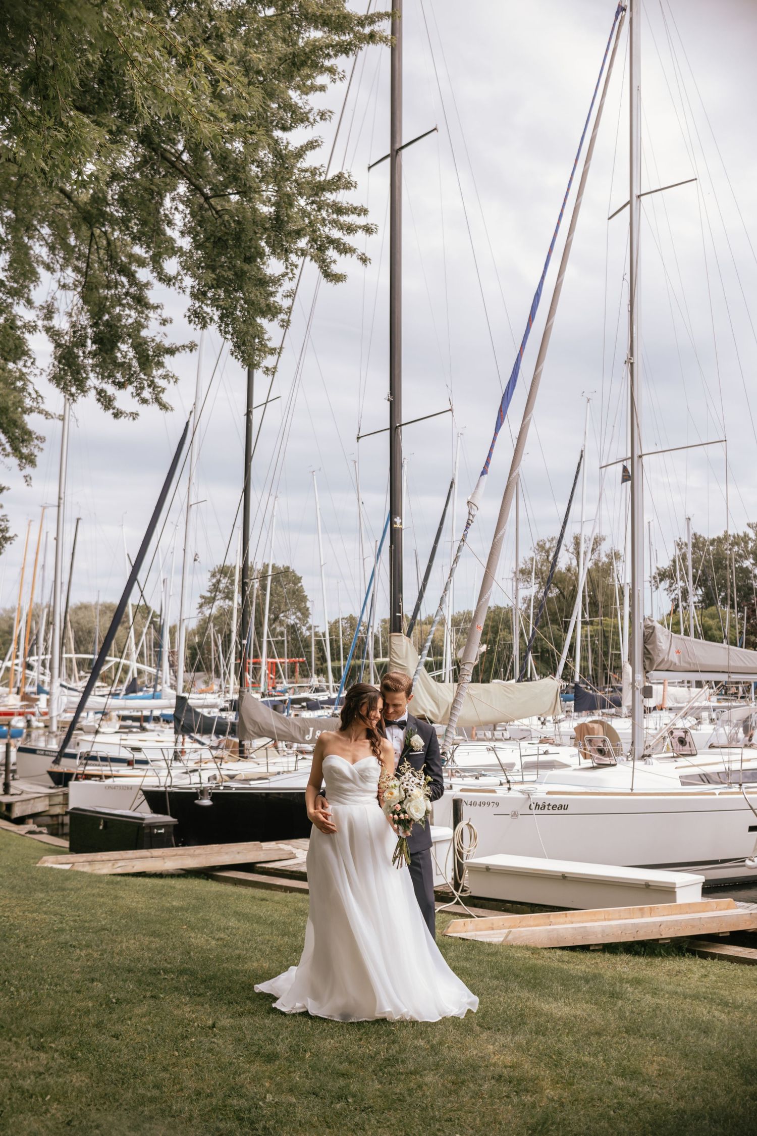 A couple poses on a grassy lawn at a marina with sailboats docked in the background on an overcast day.