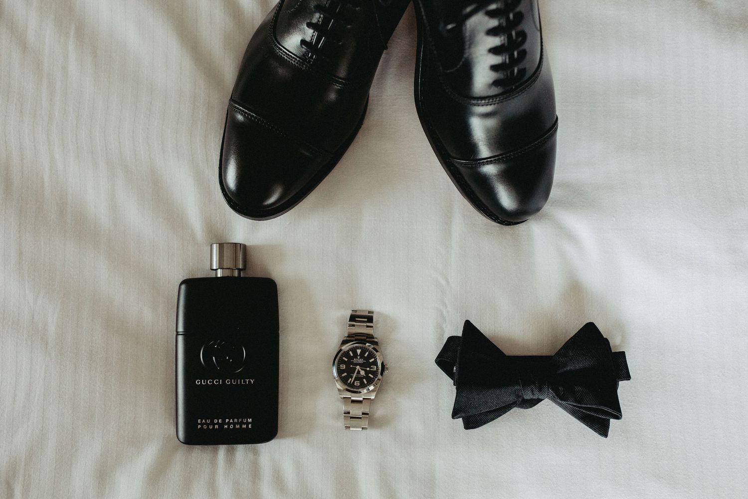 Black leather dress shoes arranged with a watch, bowtie, and cologne bottle on white fabric.