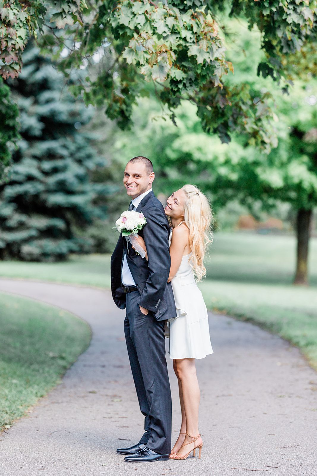A couple poses playfully on a garden path with greenery and trees in the background.