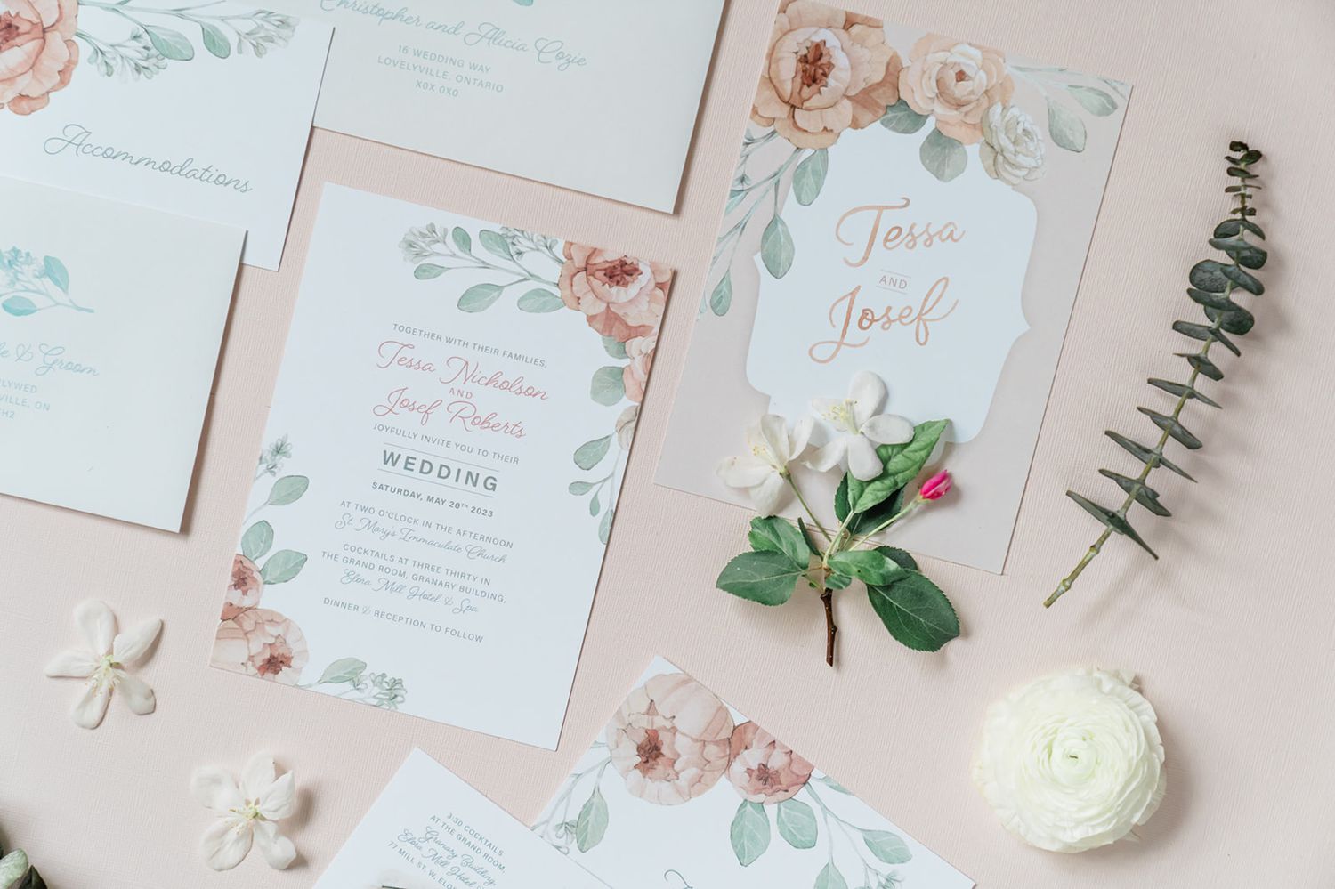 Delicate floral wedding invitations with soft pink roses and eucalyptus designs laid flat with white flower decorations.
