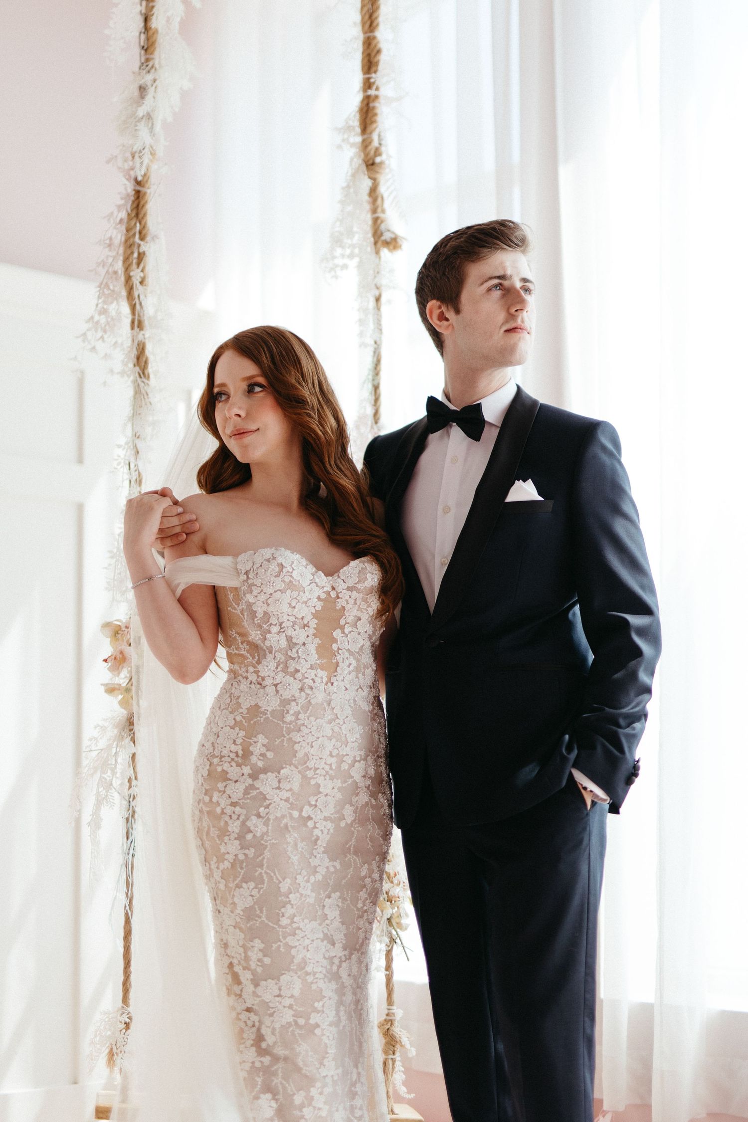 Elegant couple poses by sheer white curtains, bride in sparkly off-shoulder gown and groom in classic black tuxedo.