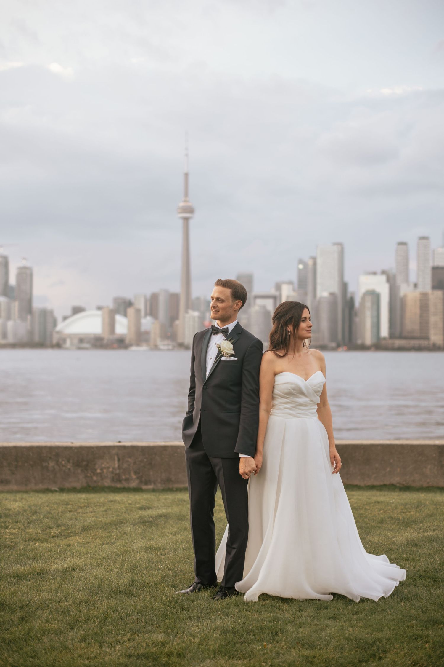 A couple in wedding attire poses along the Toronto waterfront with the iconic CN Tower and city skyline in the background.