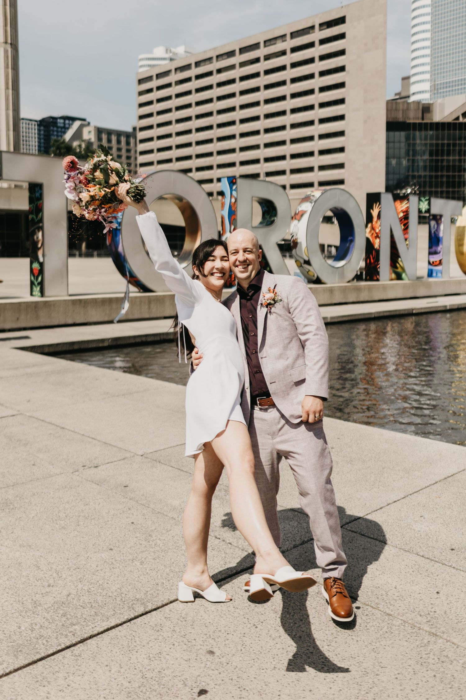 A happy couple in matching light colored outfits pose together in front of the Toronto sign on a sunny day.
