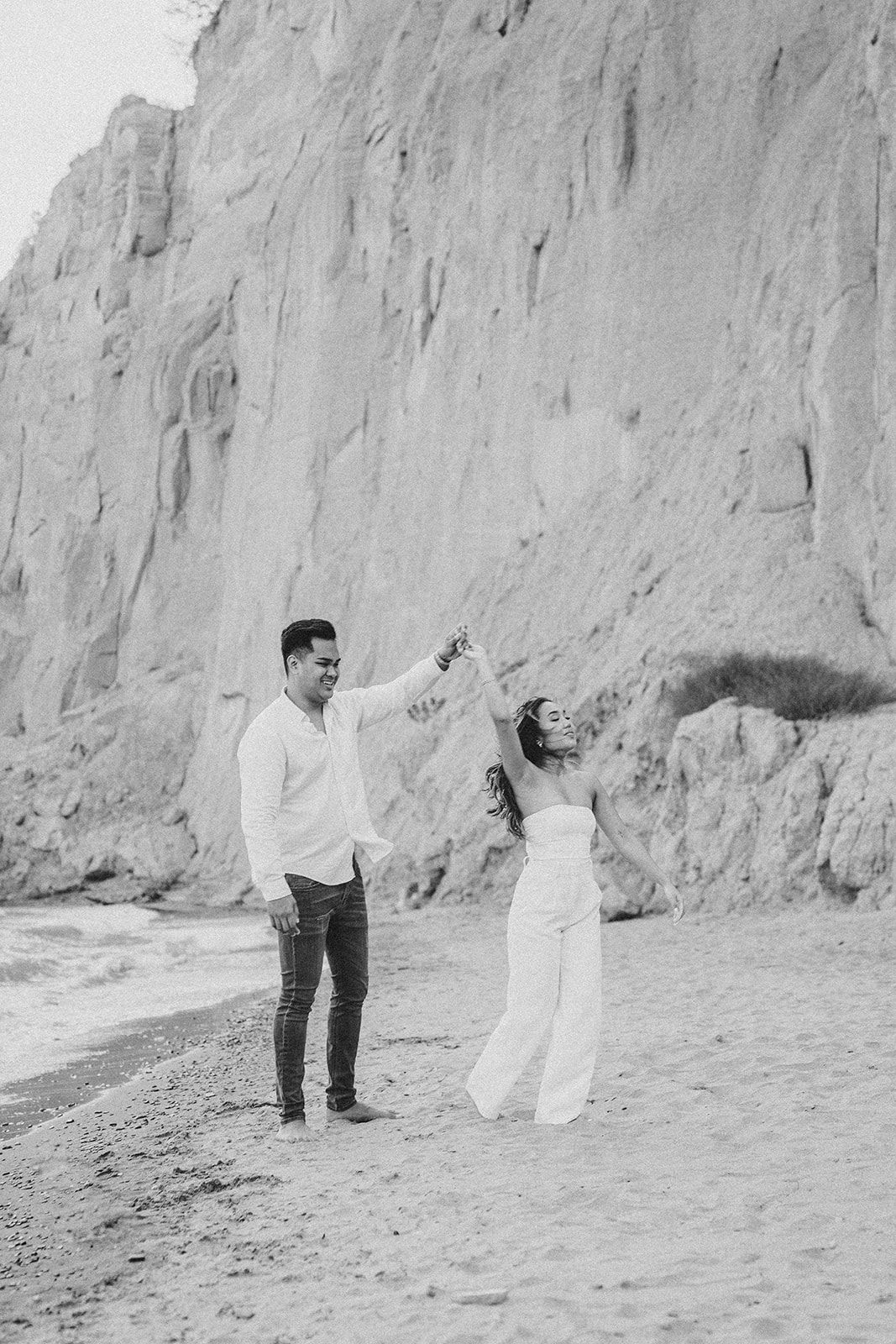 A couple dancing together on a beach with dramatic white cliffs in the background, captured in black and white.